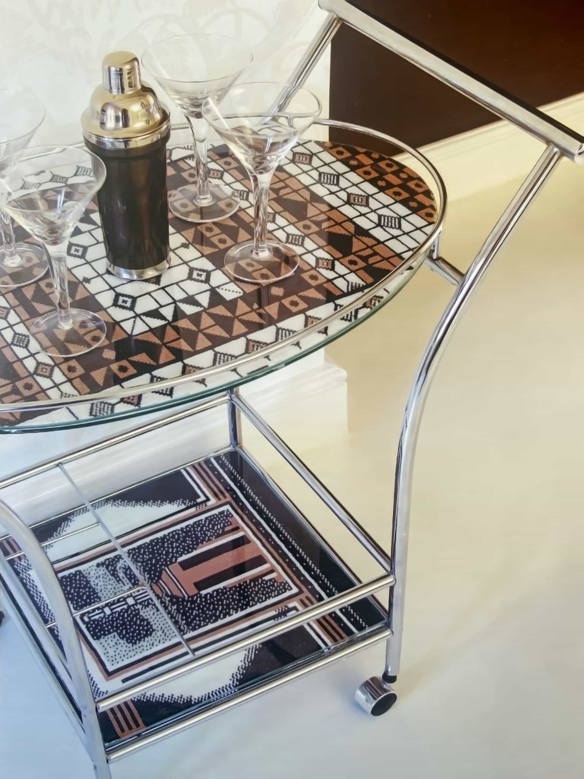 A chrome and glass drinks trolley stands on a cream floor. The trolley’s top shelf holds a silver cocktail shaker and four martini glasses on a geometric-patterned tray. Below, the lower shelf displays another pattern-covered tray with two chocolate bars.