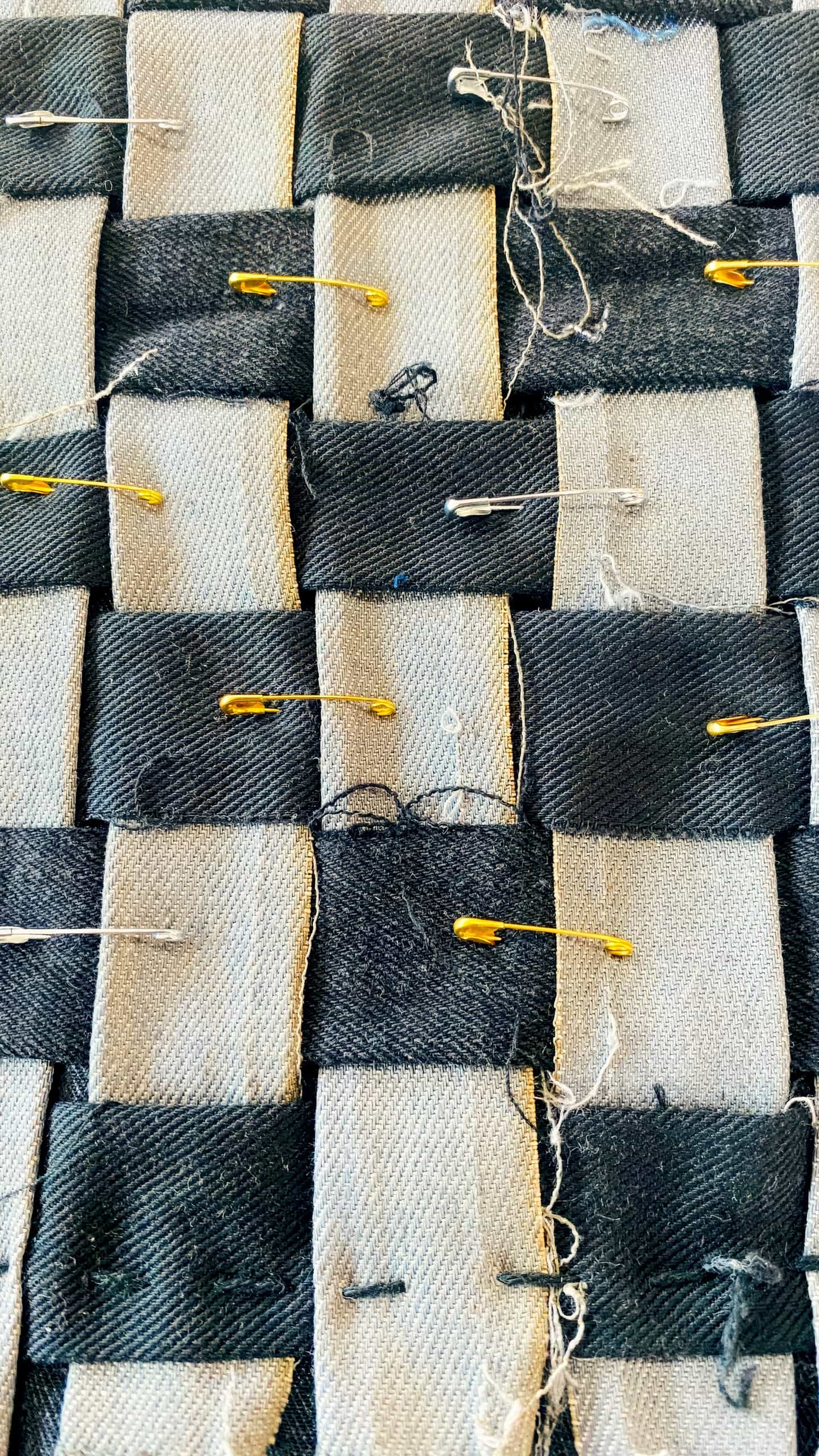 Close-up of woven black and light grey fabric strips with loose threads. Several gold and silver safety pins fasten the strips together. The woven pattern and rough textile texture are clearly visible in the image.