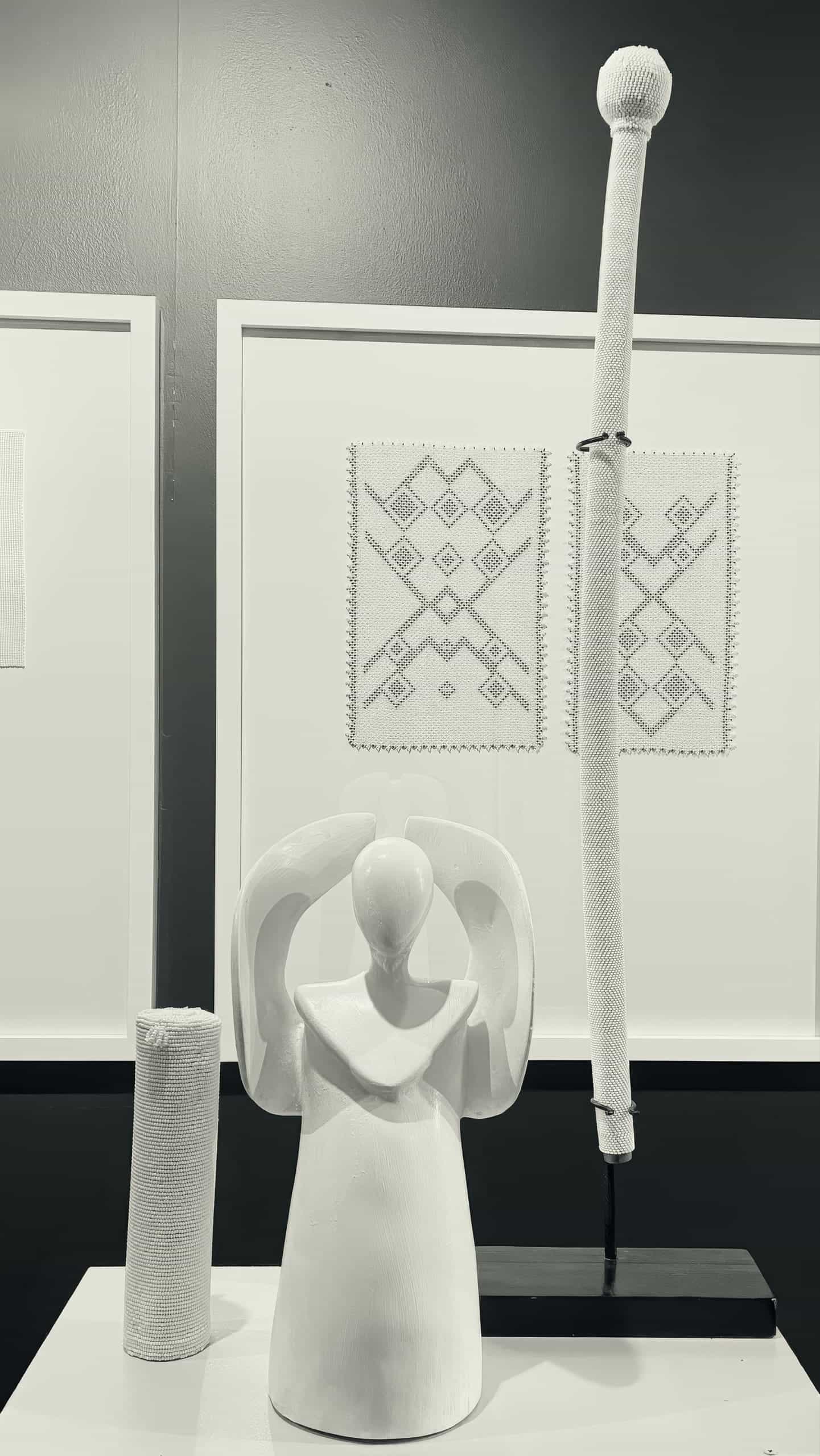 A black-and-white photo shows a smooth, abstract angel sculpture with arms raised. Behind it are two geometric embroidered fabric pieces framed on the wall, and to the right stands a tall, wrapped staff with a textured sphere on top.