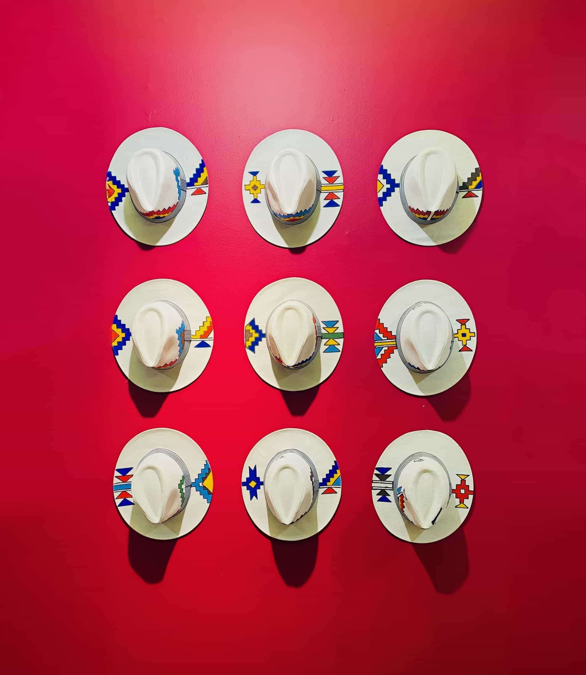 Nine white cowboy hats with colourful beaded hatbands are mounted in a three-by-three grid on a vivid red wall. Each hat sits on a round white plate, which features geometric patterns in blue, yellow, and red.