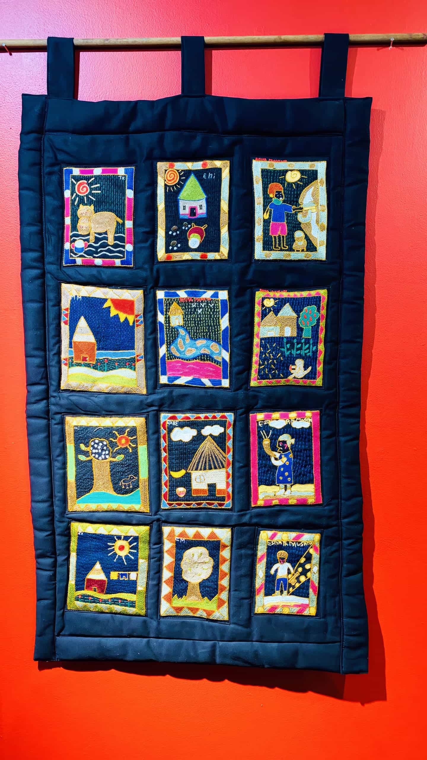 A fabric wall hanging with twelve colourful embroidered panels, each depicting different scenes and figures, bordered in yellow and blue, is set against a vibrant red wall. The panels show houses, people, trees, animals, and suns in a folk-art style.