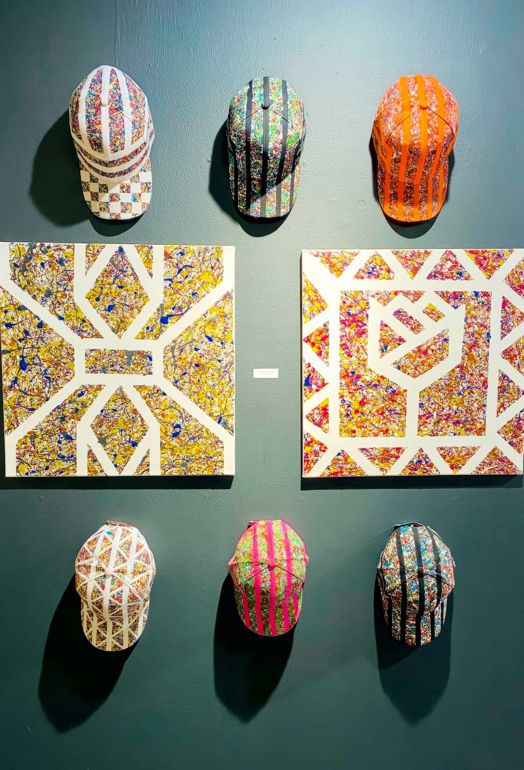 Six colourful, patterned caps are displayed on a dark wall around two abstract, geometric artworks. Titled 