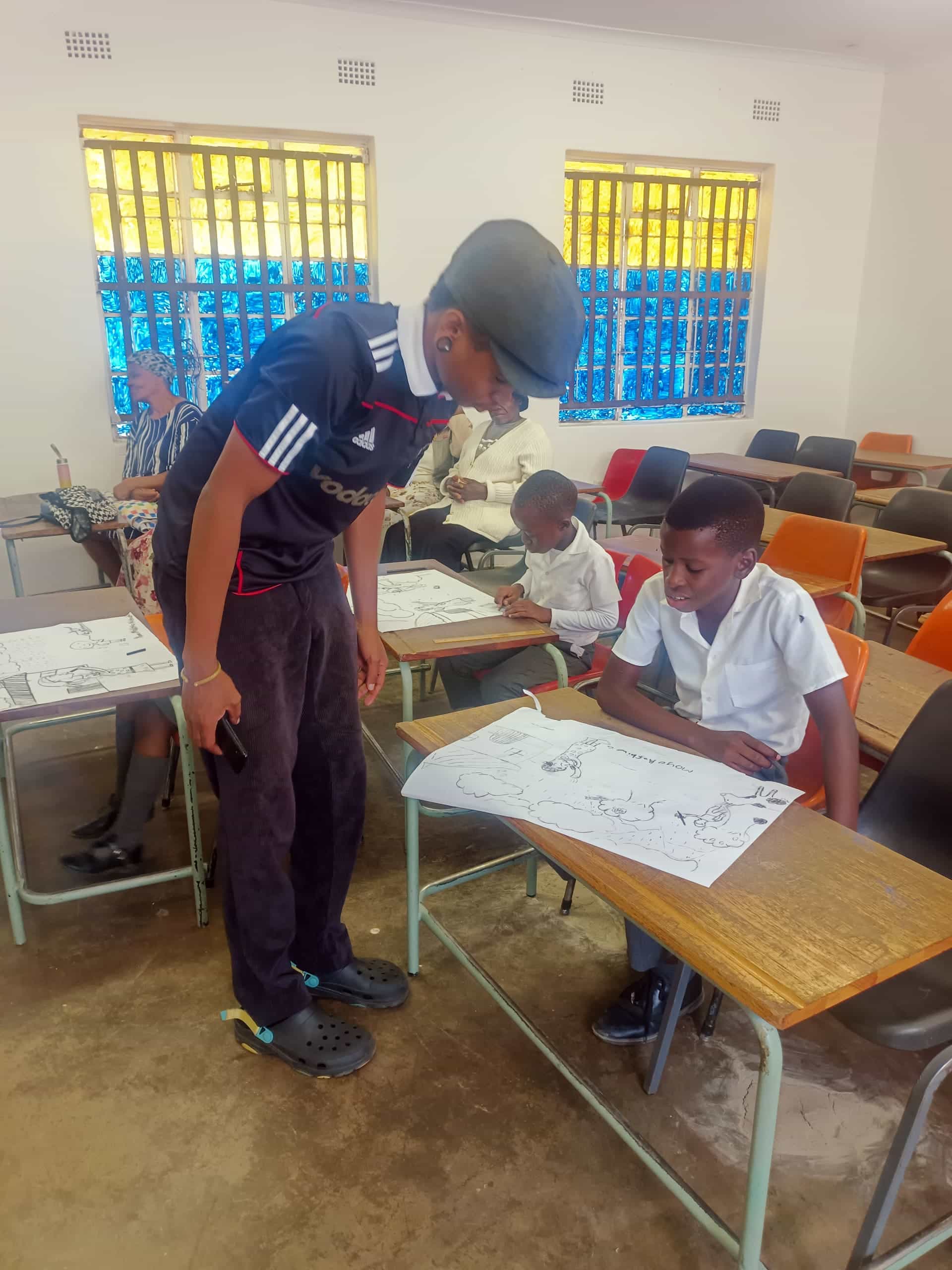 A teacher leans over to assist a pupil seated at a desk in a classroom. Both are focused on a large sheet of paper with drawings. Other pupils, also working on papers, sit at desks nearby. The classroom has bright yellow and blue window panes and scattered empty chairs.