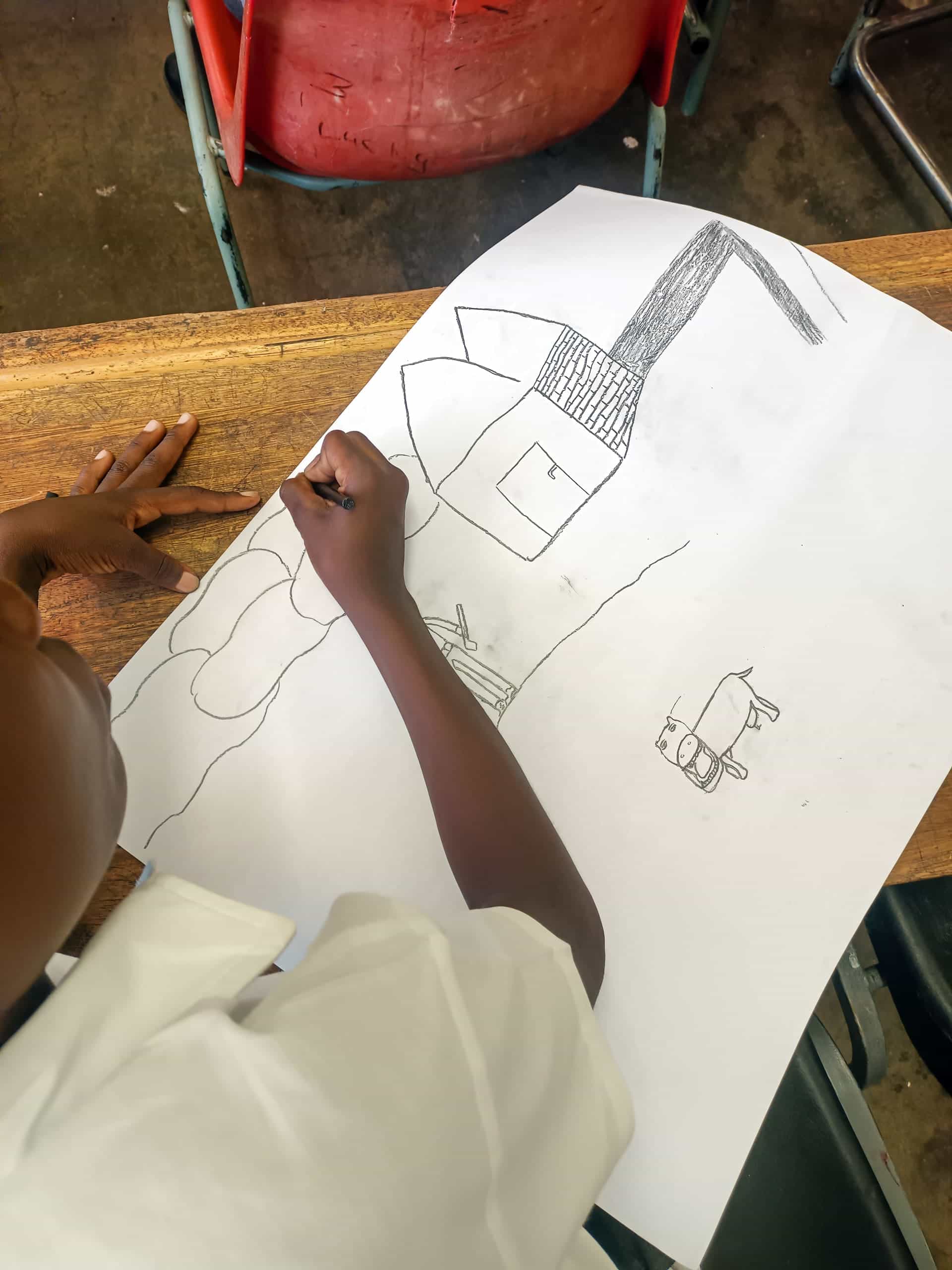 A child in a white shirt draws with a pencil on a large sheet of paper at a wooden desk. The drawing features shapes resembling a house, a tree, and a car. The child’s left hand holds the paper steady whilst they draw with their right hand.