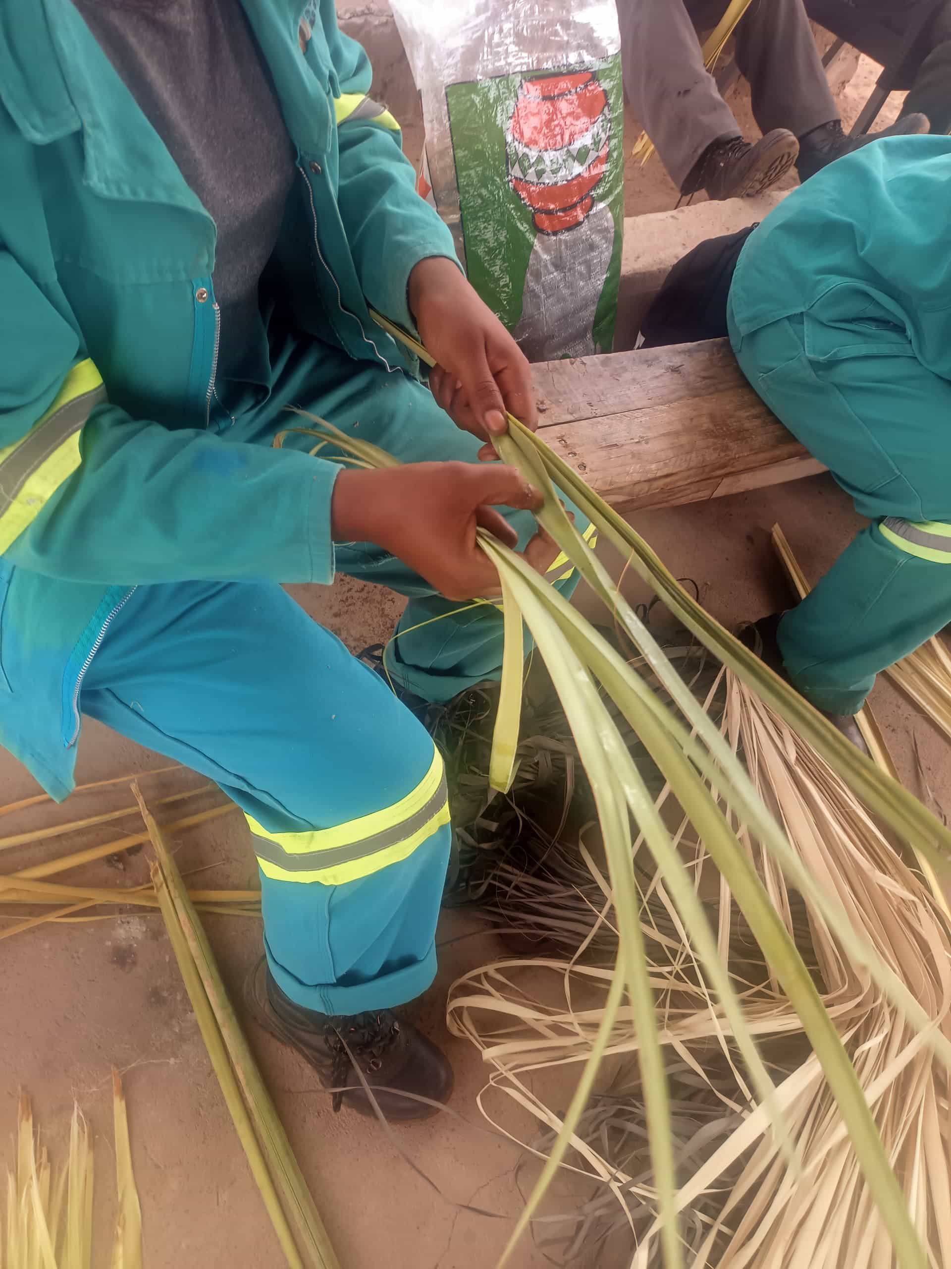 A person wearing teal work overalls and black boots weaves long, green and dry palm leaves whilst seated on a wooden bench. Another person in matching attire sits nearby. A white and green bag with red print is in the background.