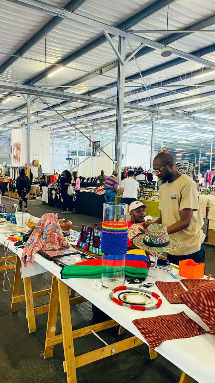 A man and woman talk at a vibrant stall with colourful handmade crafts and textiles, set inside a large indoor market. Other stalls with clothing and shoppers are visible in the spacious, industrial-style background.