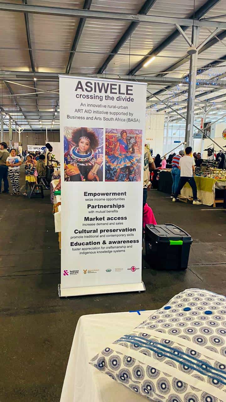 A large indoor market with people browsing stalls. In the foreground is a vertical banner for ASIWELE, a rural-urban art initiative, featuring two photos of women and text about empowerment, partnerships, and education. A patterned textile lies on a table nearby.
