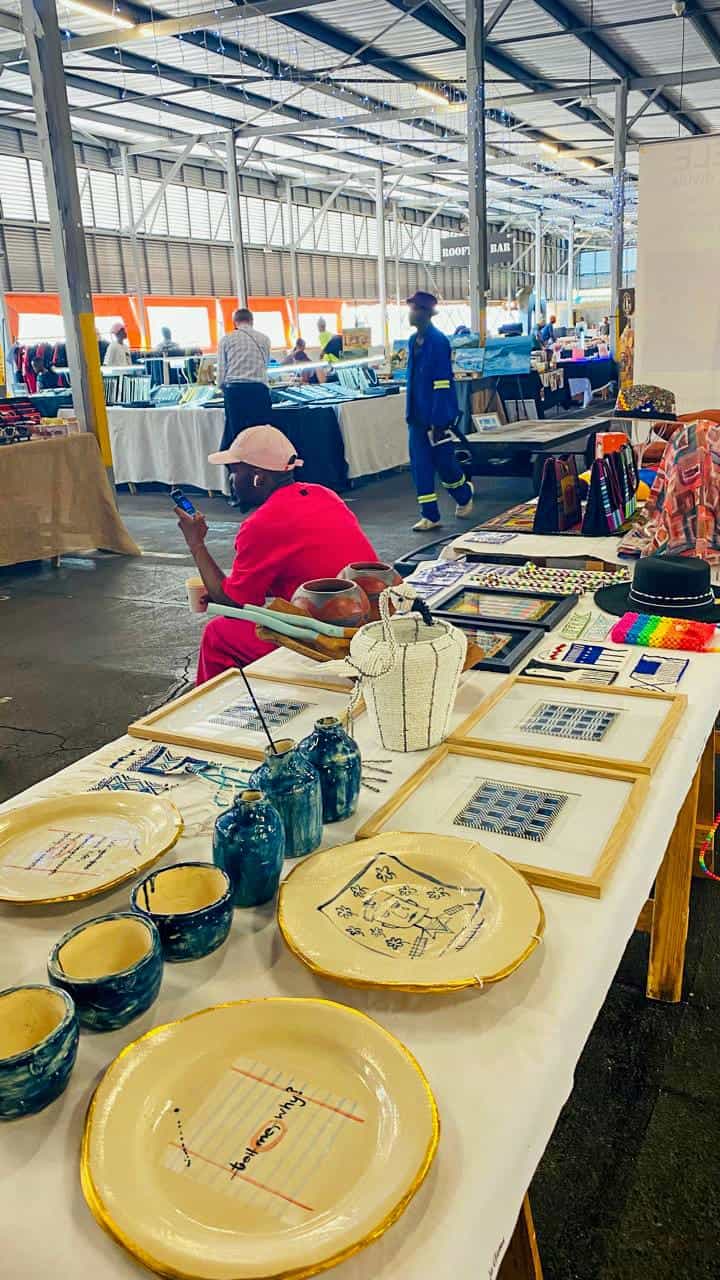 A craft market scene shows a table displaying handmade ceramics and framed art. Bowls and plates with blue patterns sit in front, with beaded jewellery and textiles further back. A person in pink sits behind the table; other shoppers browse stalls in a spacious indoor venue.