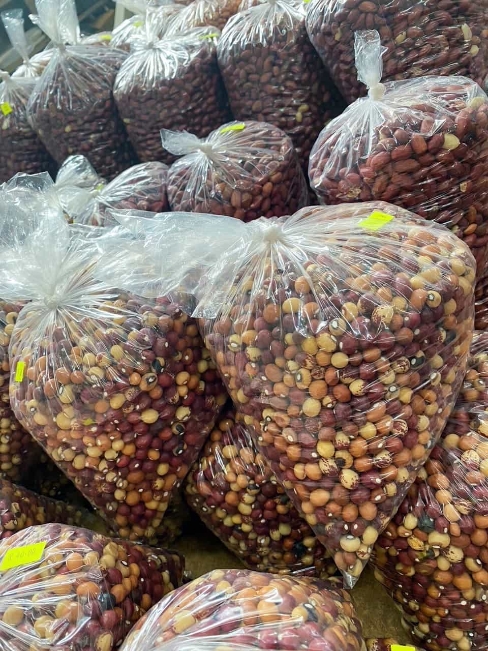 Large clear plastic bags filled with a mix of assorted dried beans and pulses are stacked in rows. The beans inside are of various colours including brown, beige, and red. Each bag has a small yellow price sticker attached. The bags are tightly packed together.