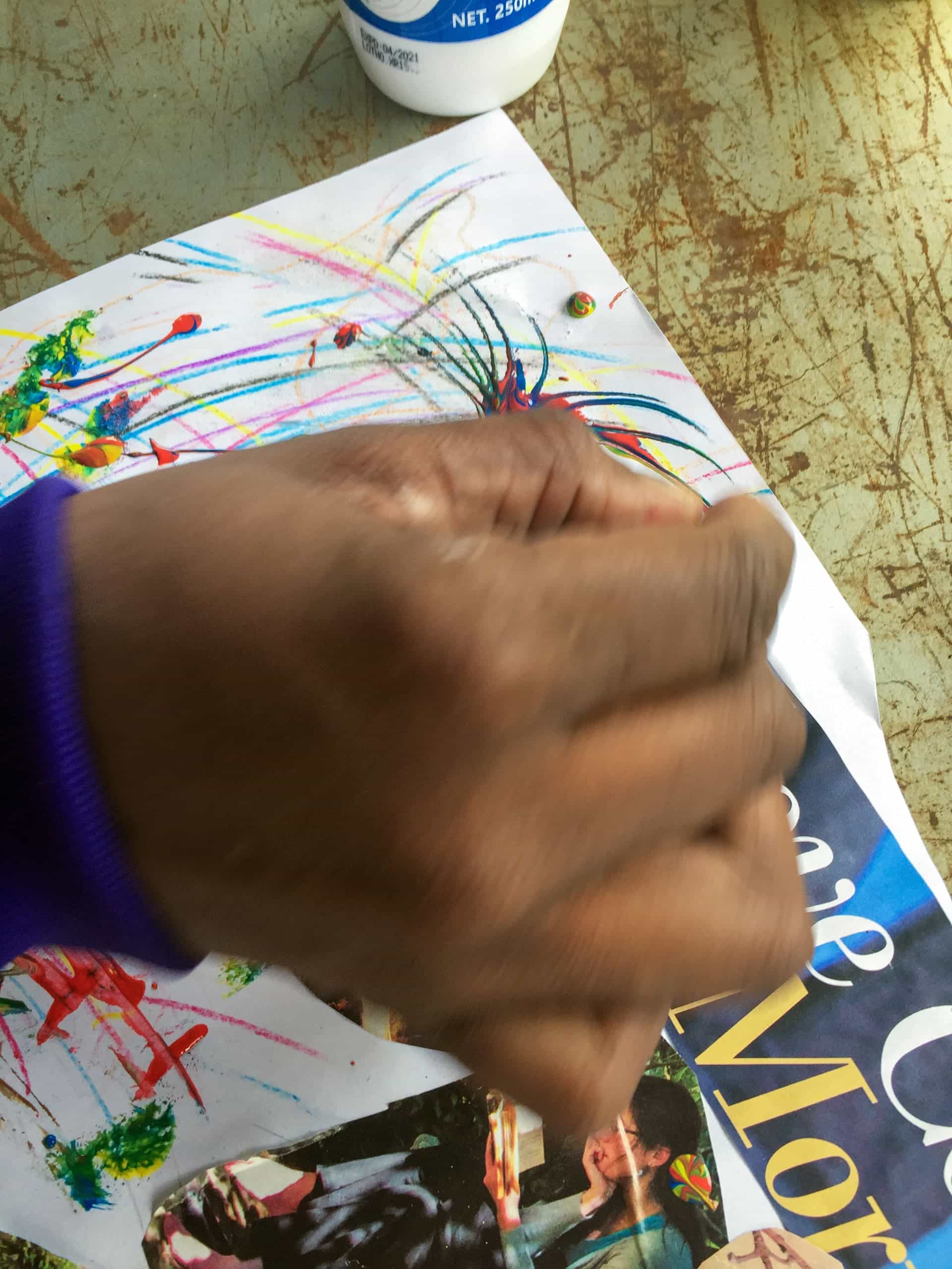 A hand in motion blurs over a piece of white paper with colourful crayon scribbles and magazine cut-outs. Part of a blue sleeve is visible. At the top, there’s a partially seen white glue bottle on a worn, greenish surface.