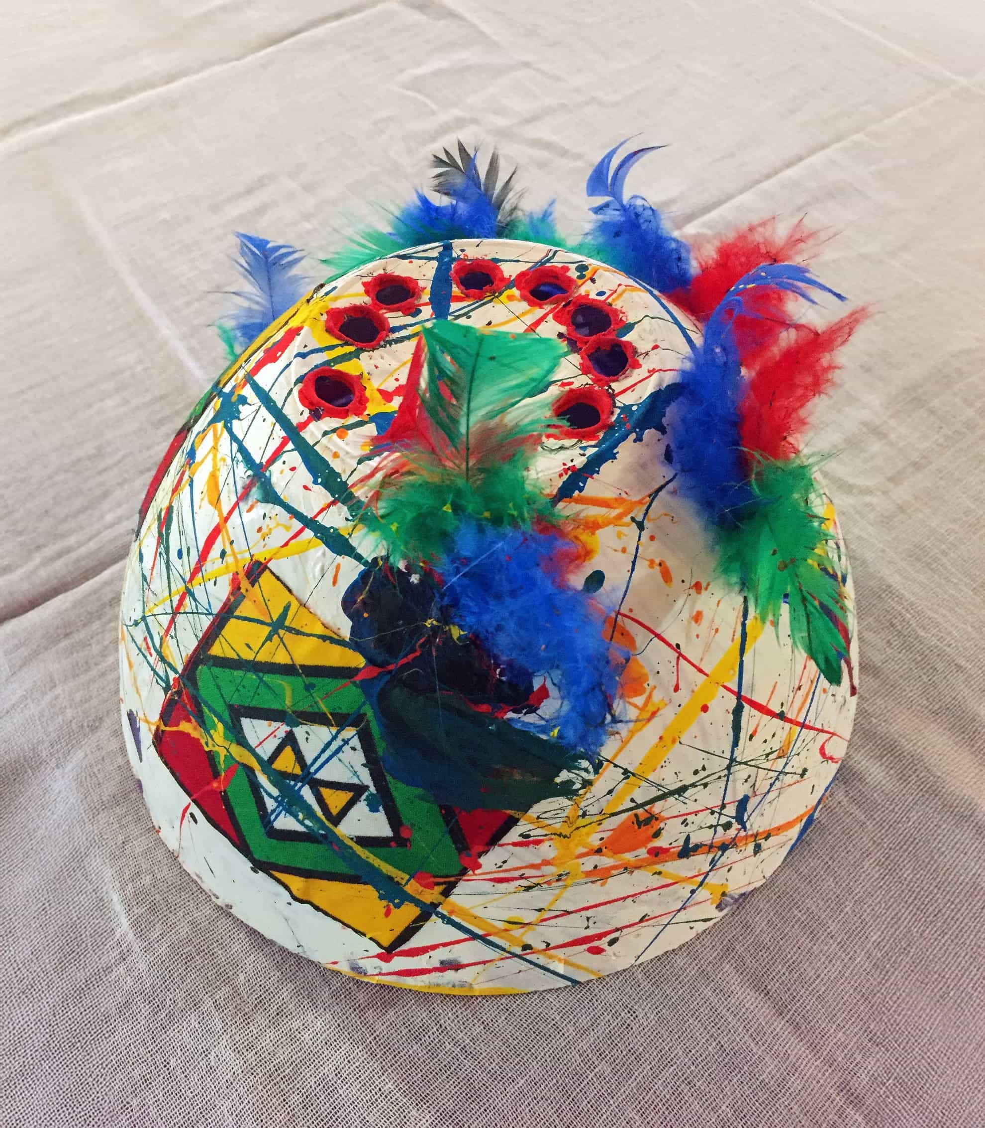 A white papier-mâché mask decorated with colourful paint splatters, geometric shapes, and circular red gems. Blue, green, and red feathers are attached on top, giving the mask a festive and artistic appearance. The mask sits on a textured cloth surface.