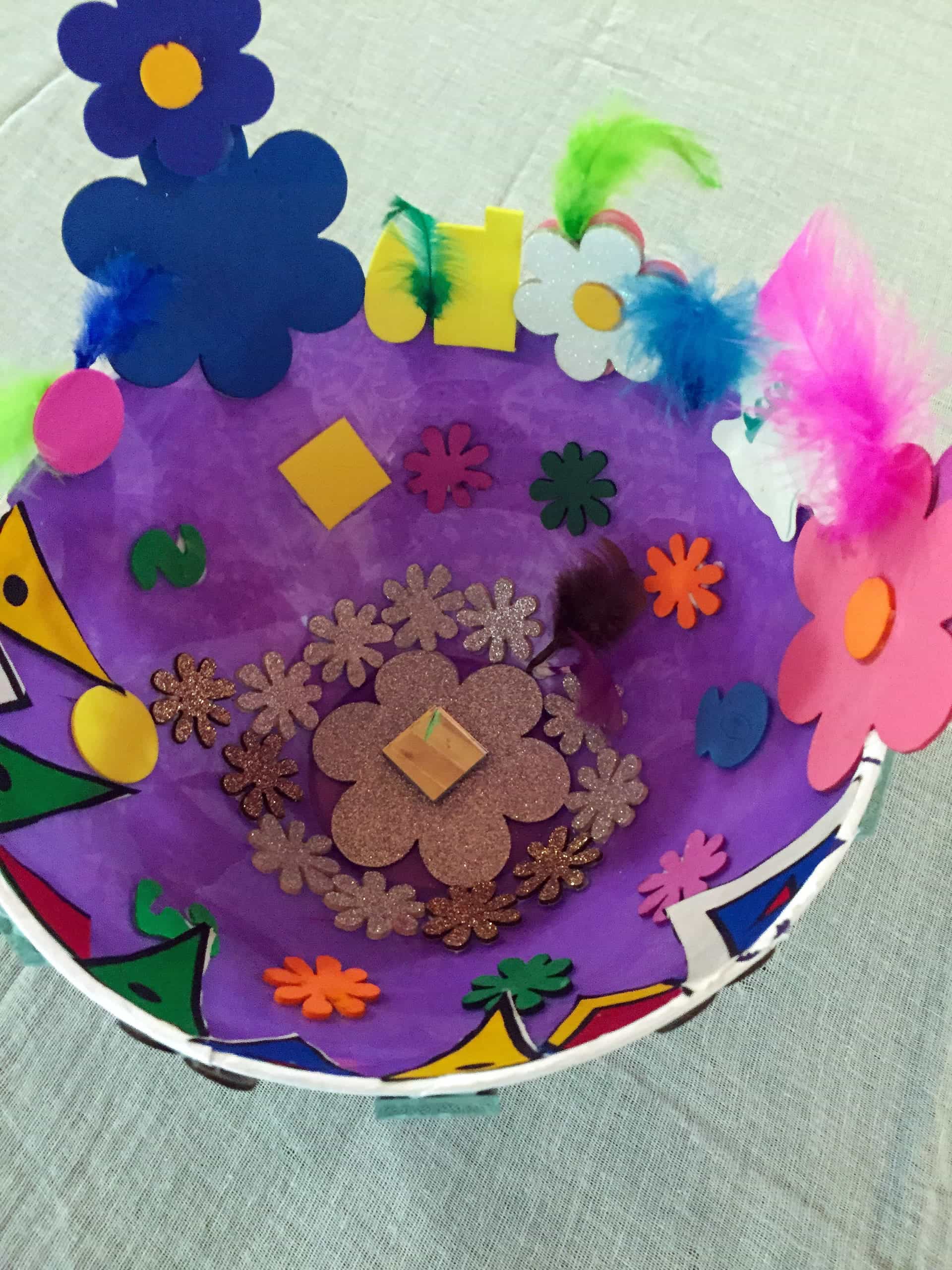 A colourful, handmade bowl decorated with large foam flowers, glittery flower shapes, feathers, and small foam shapes. The bowl’s interior is mostly purple, with an array of bright decorations inside and around the rim, set on a pale textured surface.