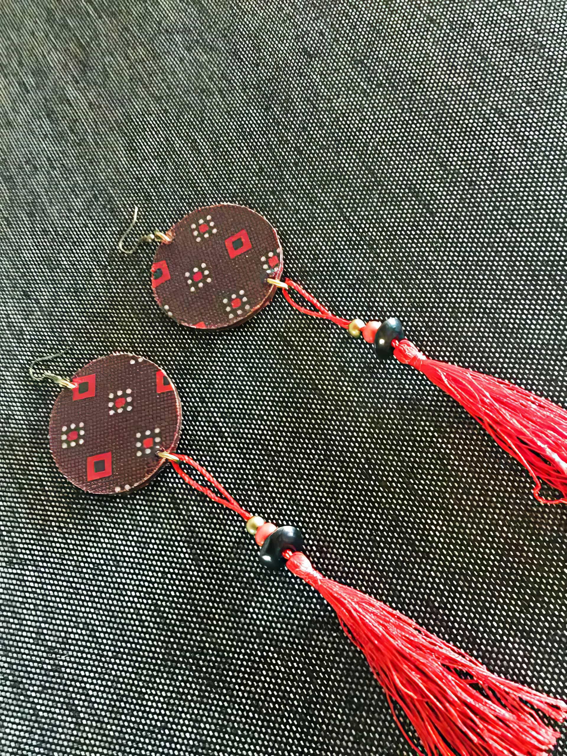 A pair of handmade earrings with round, patterned discs featuring red and white diamond shapes, black beads, gold accents, and long, bright red tassels, displayed on a textured dark grey fabric background.