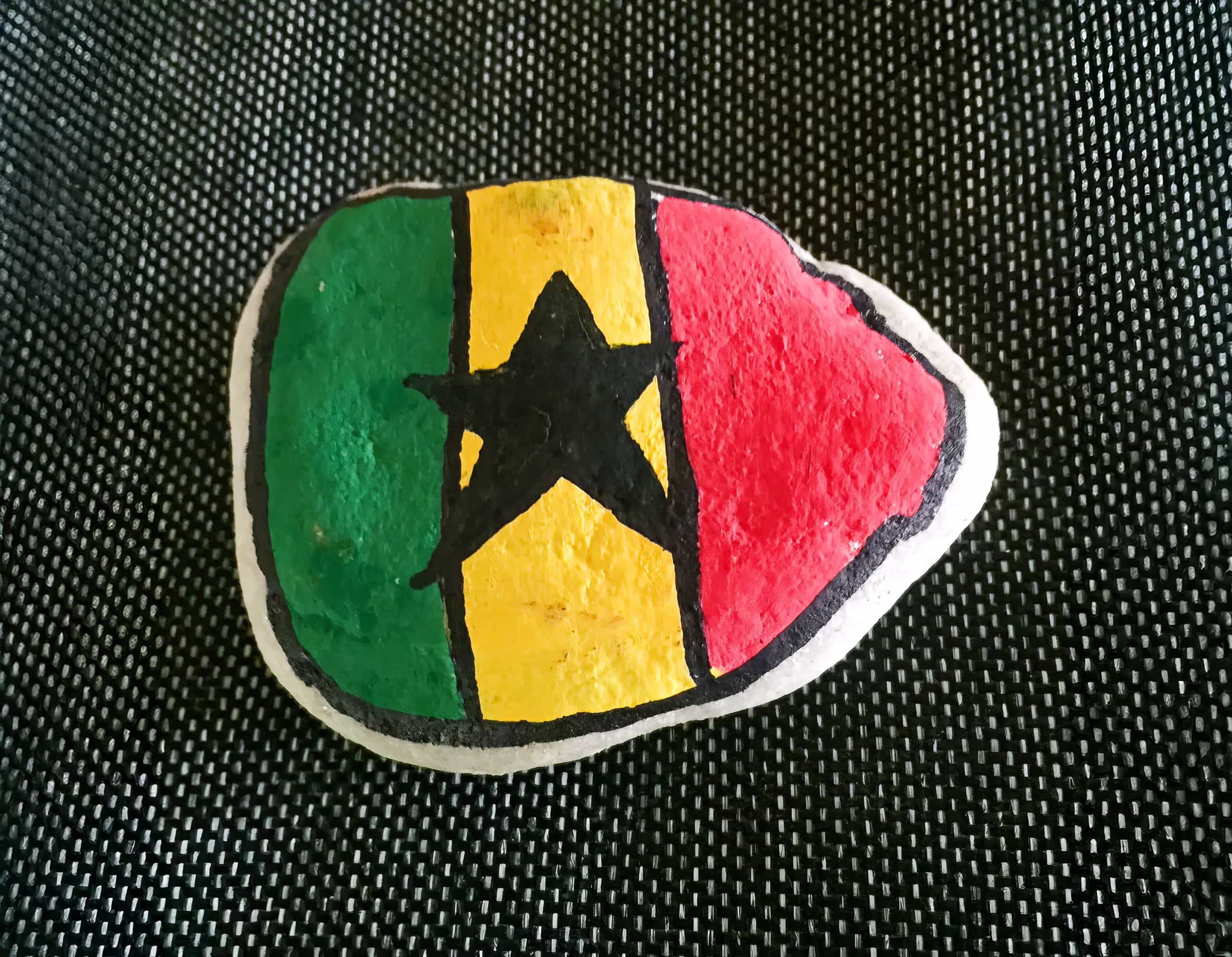 A stone painted with the Ghana flag: vertical green, yellow, and red stripes, with a black star in the centre of the yellow stripe. The stone has a black outline and rests on a textured, dark background.
