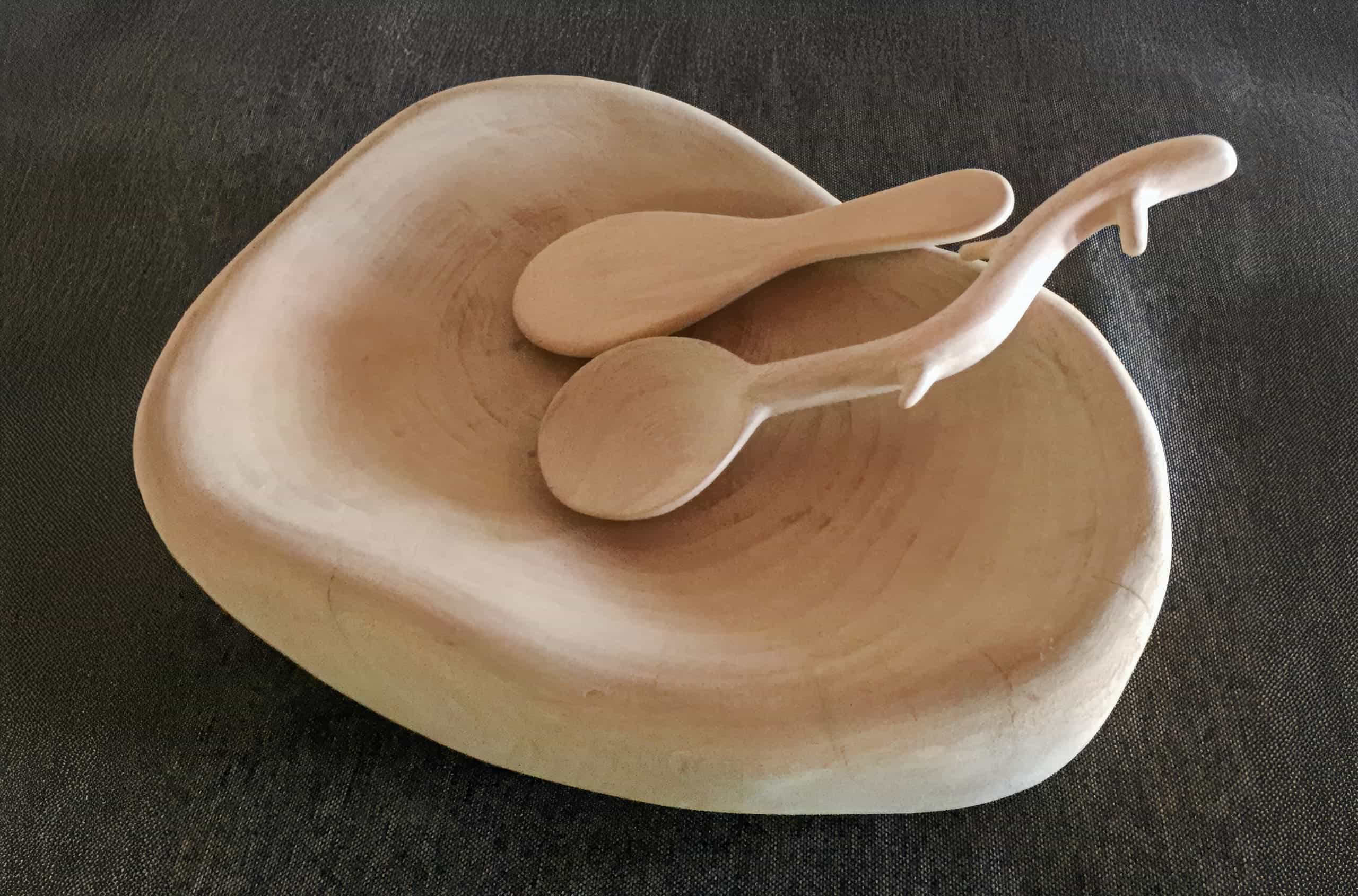 A smooth, light-coloured wooden bowl with an organic, asymmetrical shape sits on a dark textured surface. Two matching wooden utensils rest inside, one of which has a unique handle resembling an animal or branch with protruding elements.
