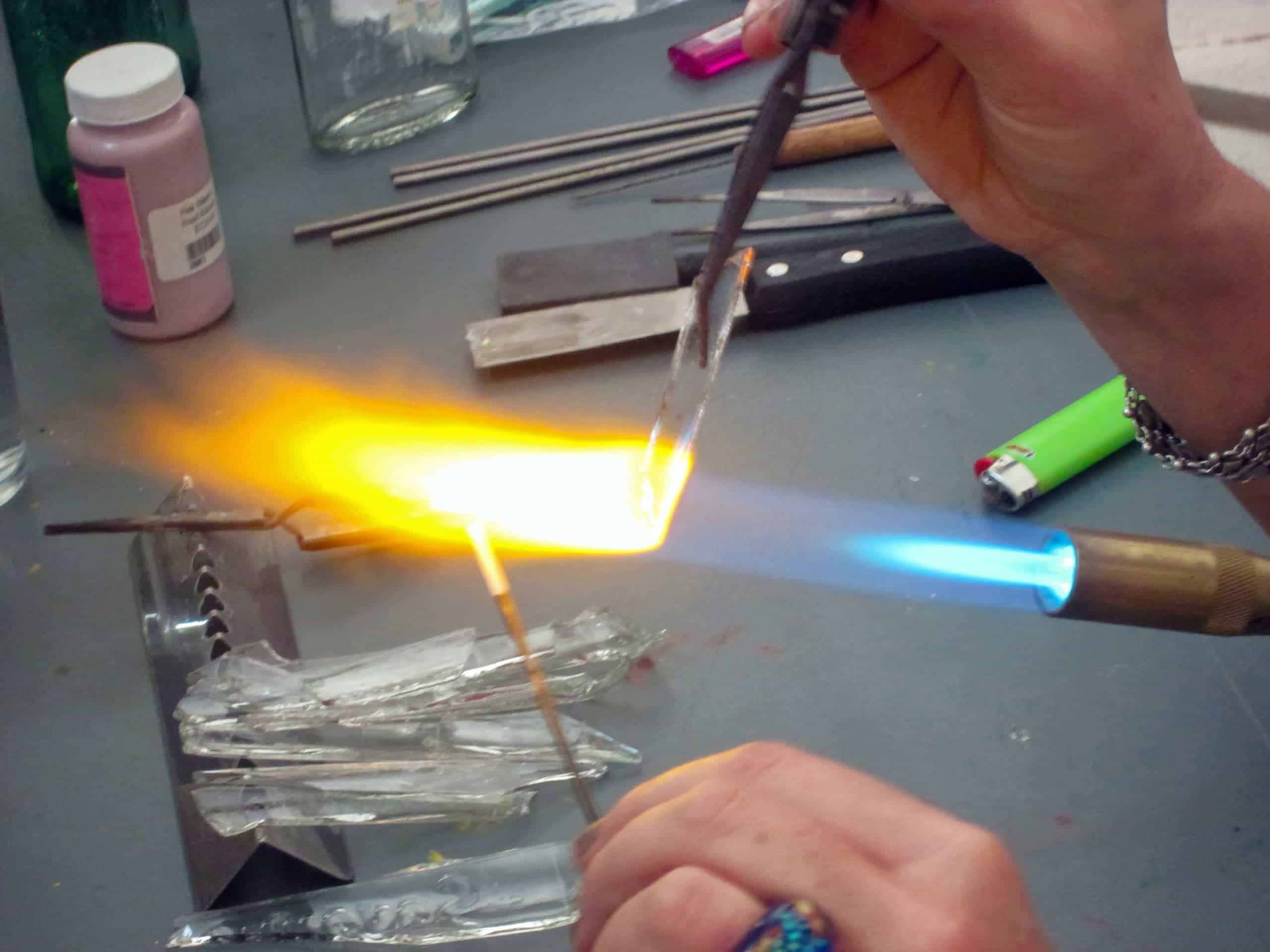 A person uses a handheld blowtorch to heat and shape clear glass rods, producing a bright orange-yellow flame. Various glass fragments, metal tools, a lighter, and jars are scattered on the work surface. Only the person's hands are visible.