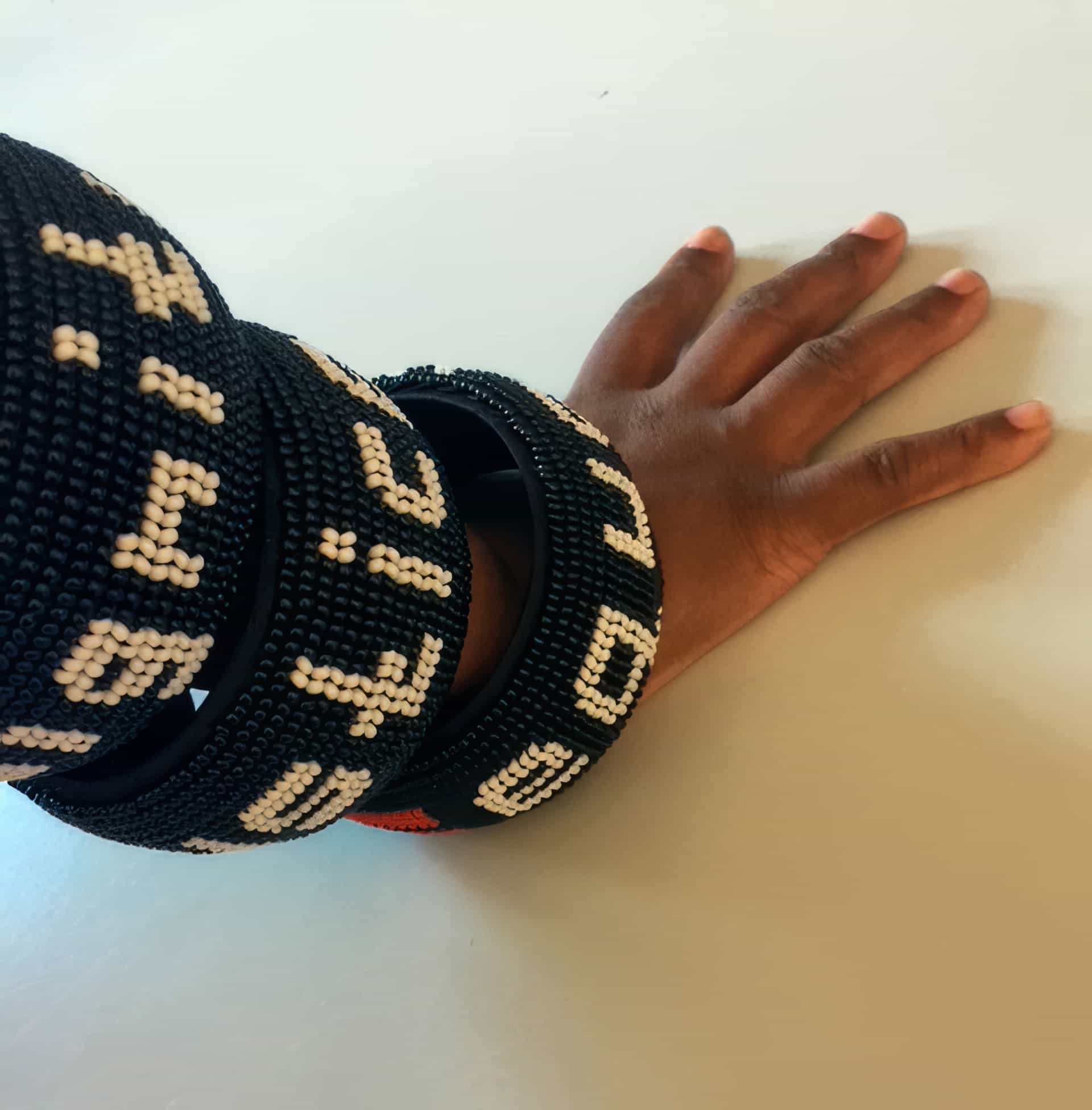 A hand with medium-brown skin rests on a white surface, wearing three large, black beaded bracelets with white geometric patterns and text-like symbols. The bracelets appear chunky and are stacked on the wrist and forearm.
