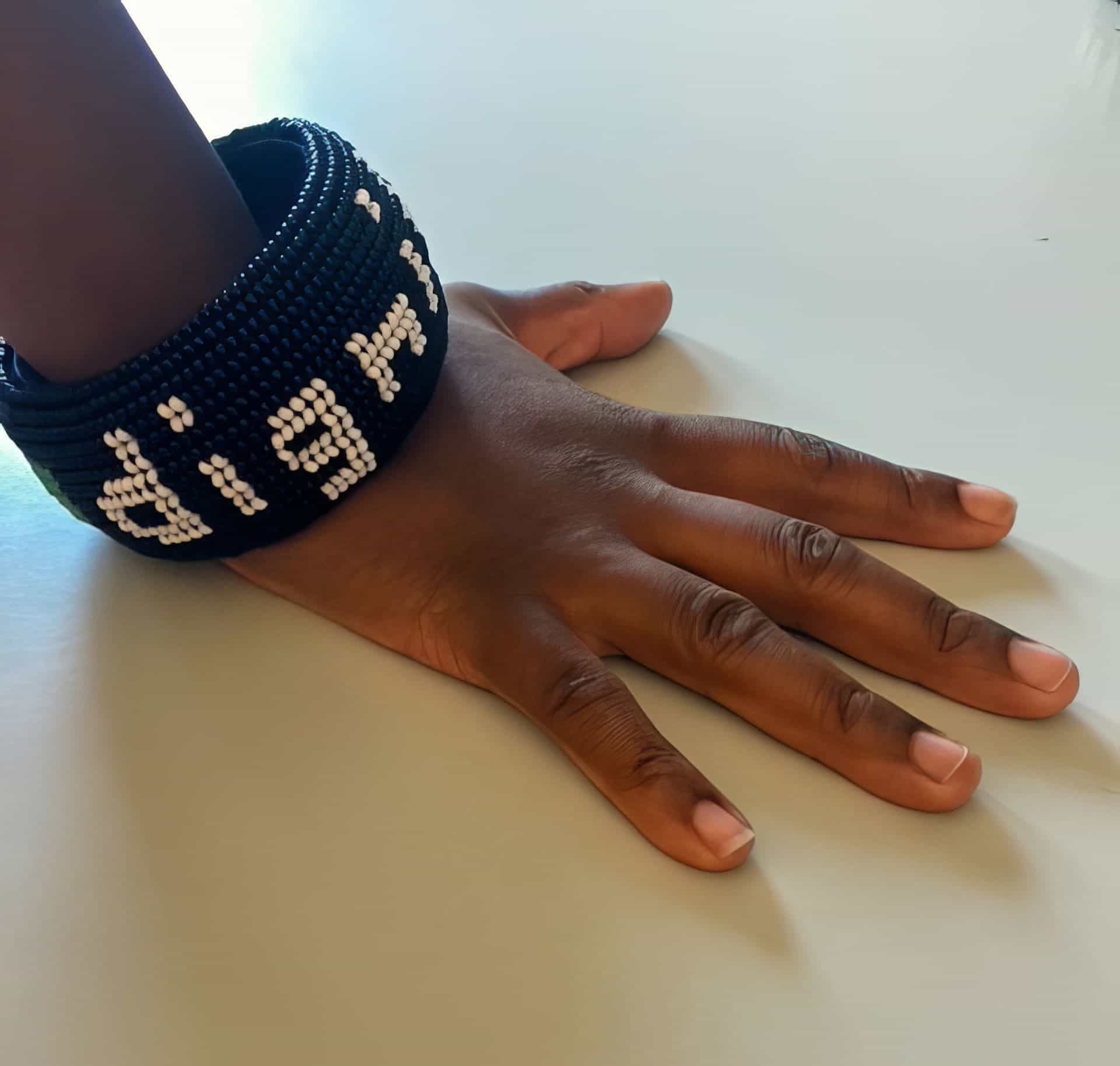 A hand with medium brown skin rests on a white surface, wearing two wide, black beaded bracelets with white beaded letters spelling “dignity.” The lighting is soft, with the bracelets as the focal point.