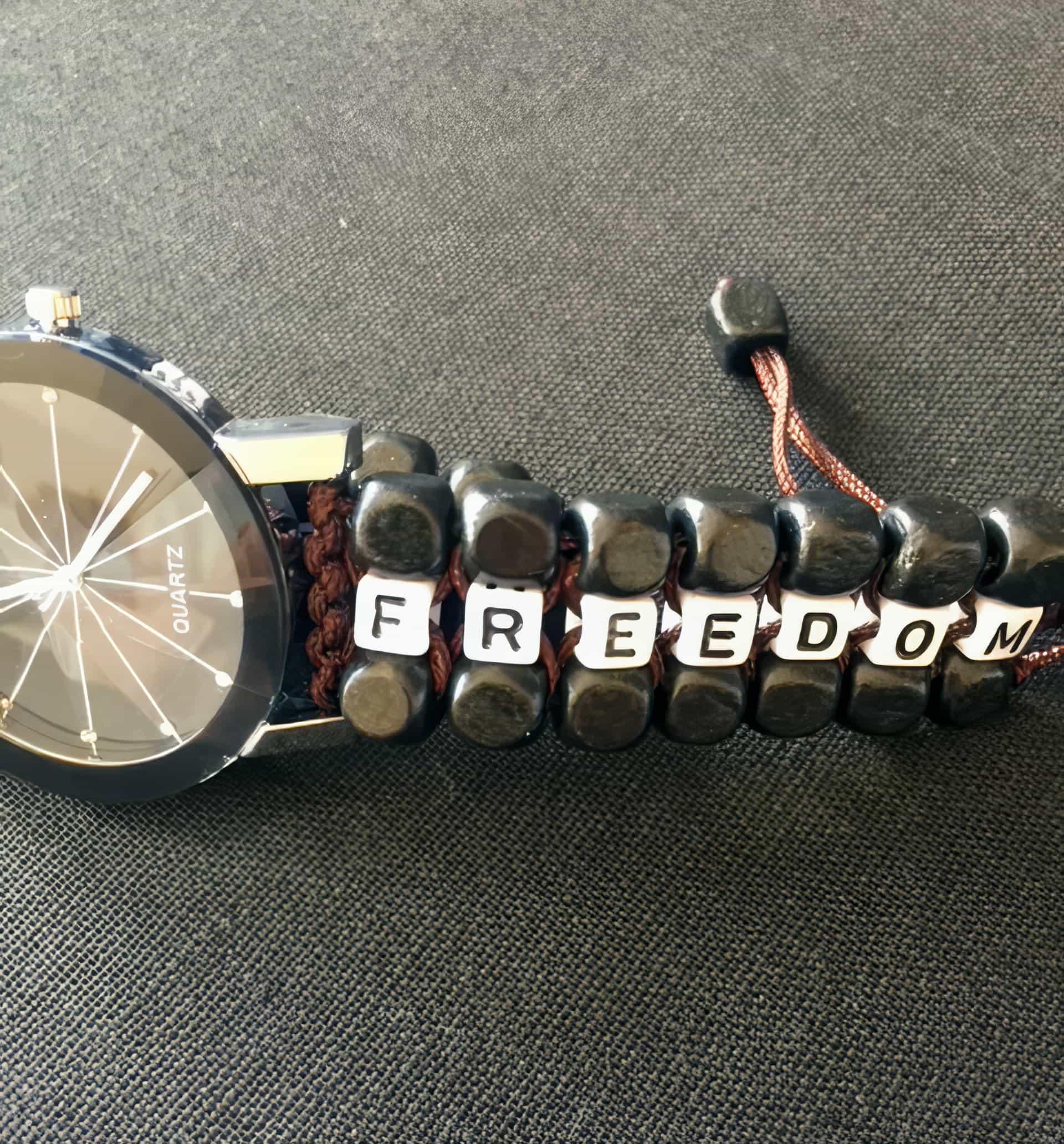 A black wristwatch with a shiny face lies on a dark fabric. The strap is made of dark beads and brown string, with white cube beads spelling “FREEDOM” in black letters woven into the strap.