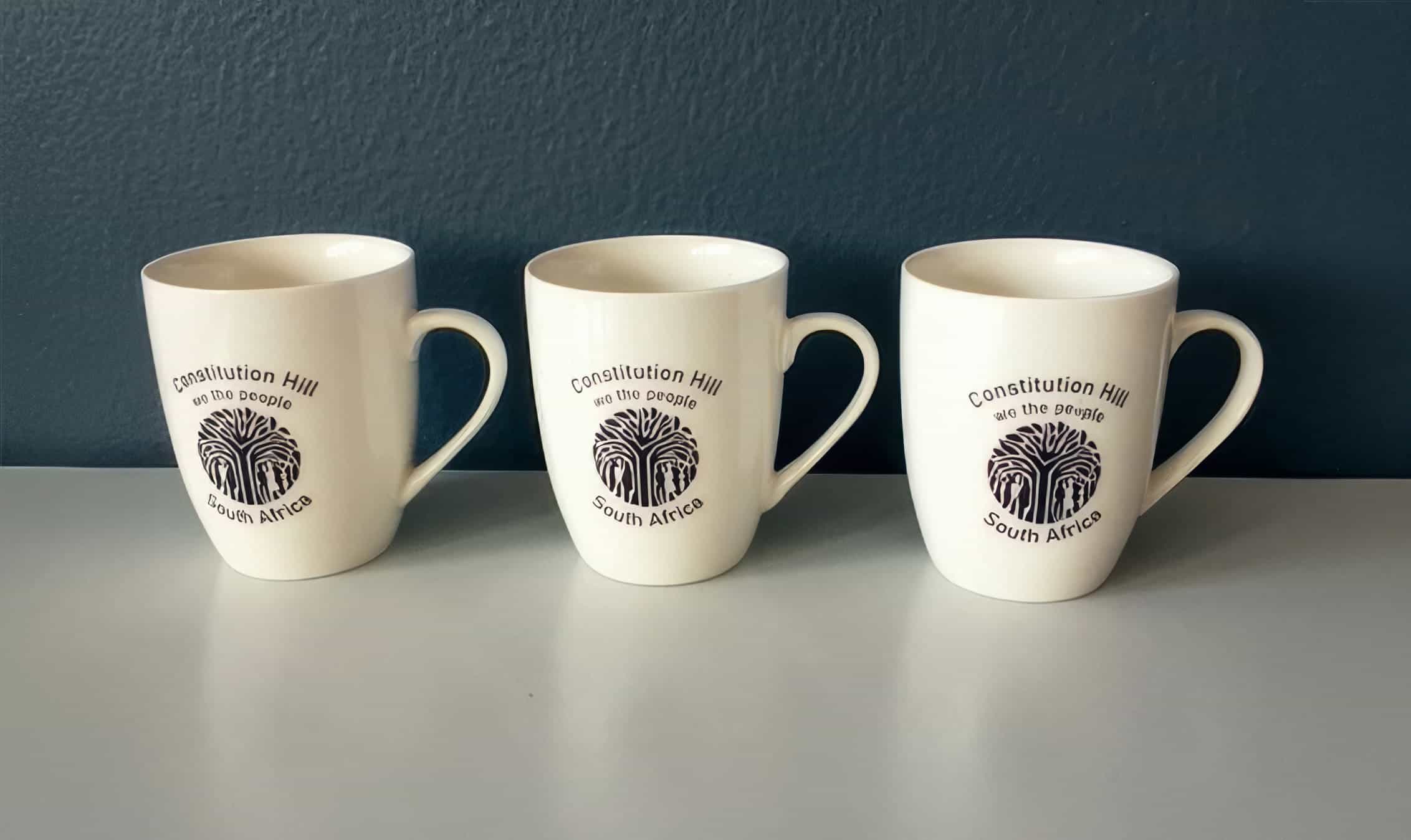 Three white ceramic mugs sit side by side on a light grey surface against a dark blue wall. Each mug has a handle and features the same black logo and text: “Constitution Hill, We the People, South Africa” with a stylised design below the text.
