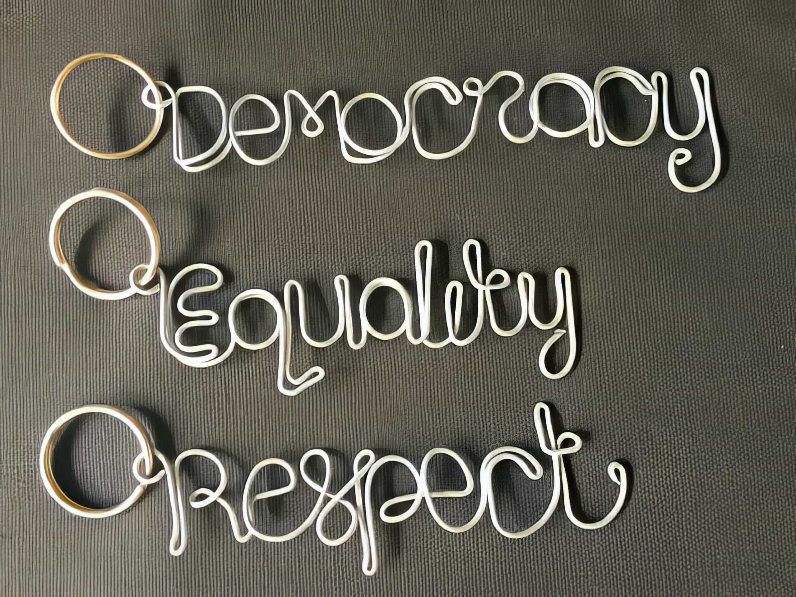 Three keyrings shaped from wire spell out “Democracy,” “Equality,” and “Respect” in cursive writing. The words are arranged one below the other on a textured dark background. The wire is light-coloured, standing out against the surface.