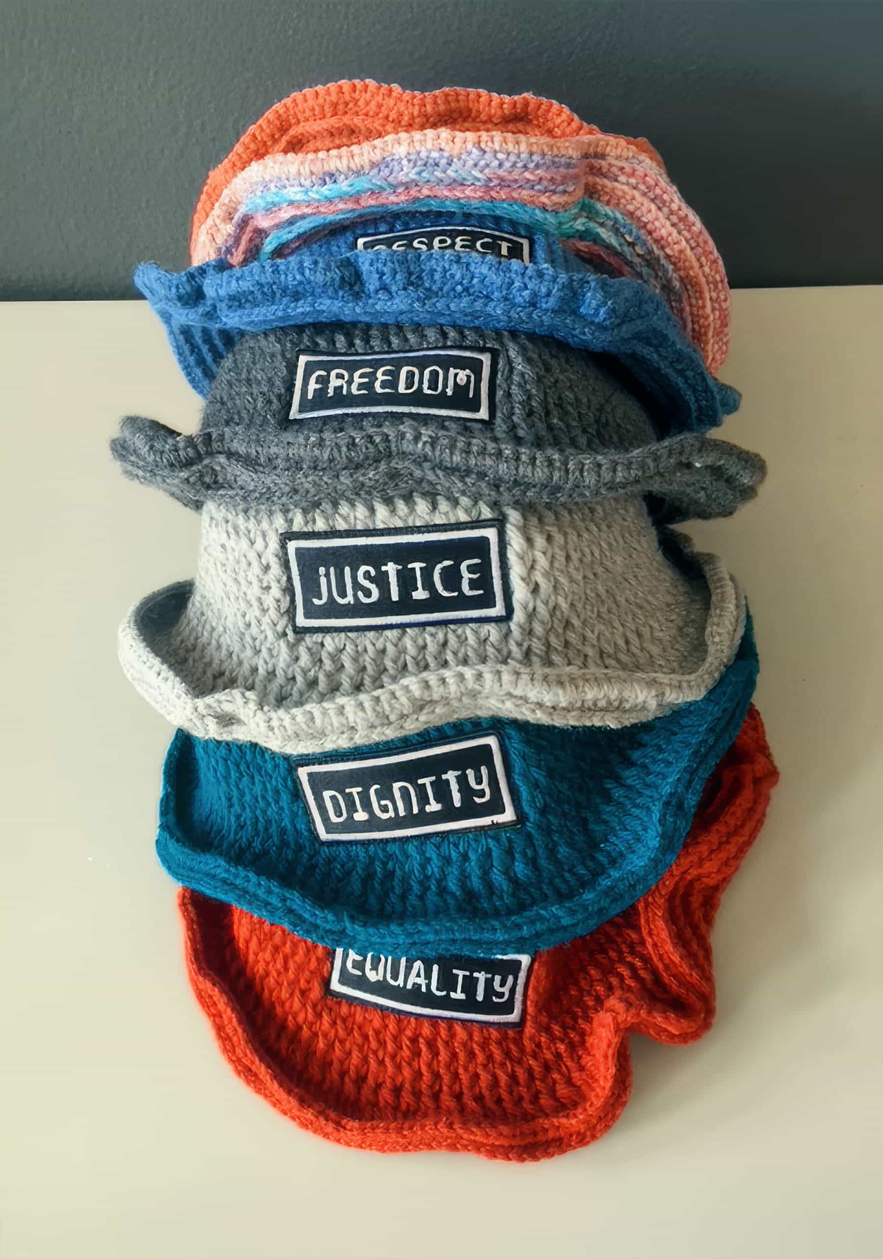 A stack of five crocheted hats sits on a white surface. Each hat features a patch with a different word: 