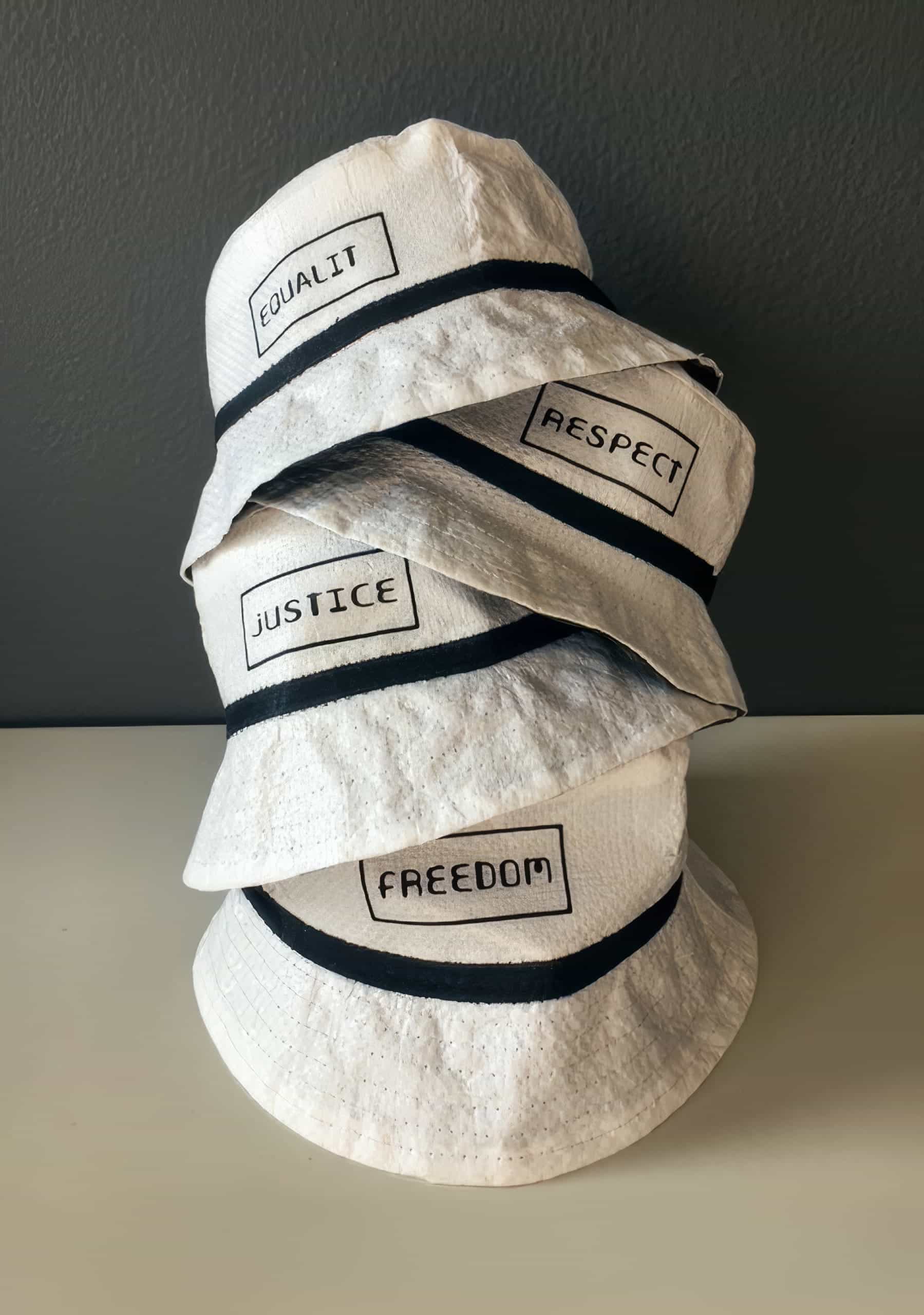 Four white bucket hats are stacked on a white surface against a dark wall. Each hat has a black band and a rectangular patch with black text: 