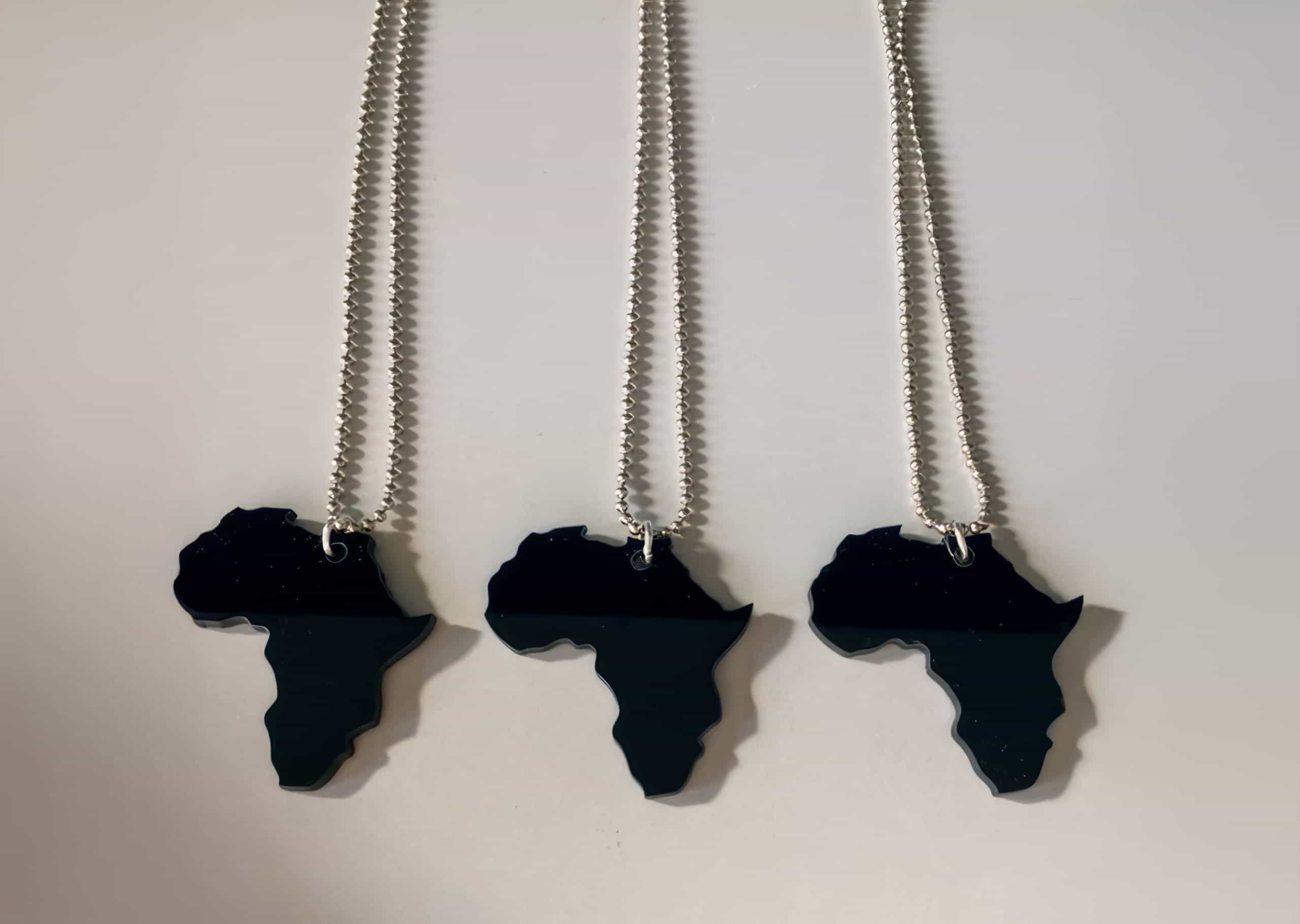 Three silver chains with black pendants shaped like the African continent are laid out vertically on a white surface, evenly spaced and parallel to each other. The pendants are glossy and reflect light, highlighting their distinct Africa silhouette.