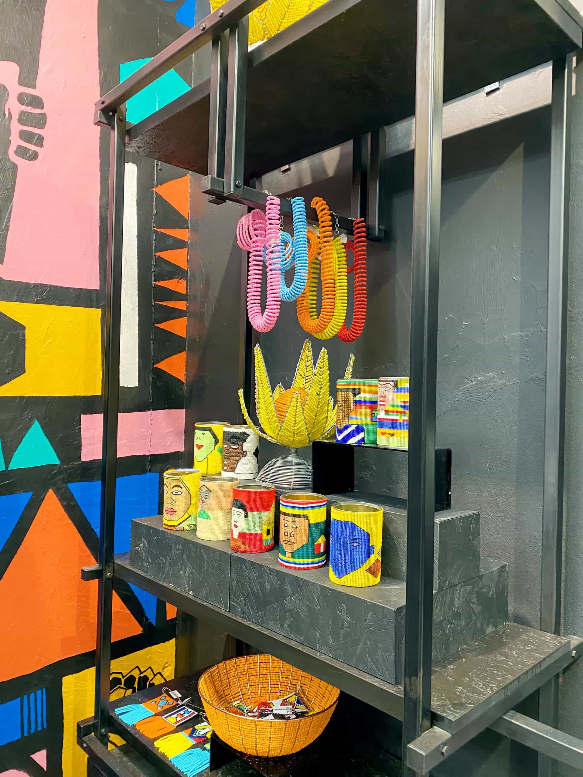 A black metal shelf displays colourful handmade crafts, including beaded tins, woven wire sculptures, and coiled necklaces. A bright geometric mural with yellow, blue, red, and black shapes decorates the wall behind the shelf.