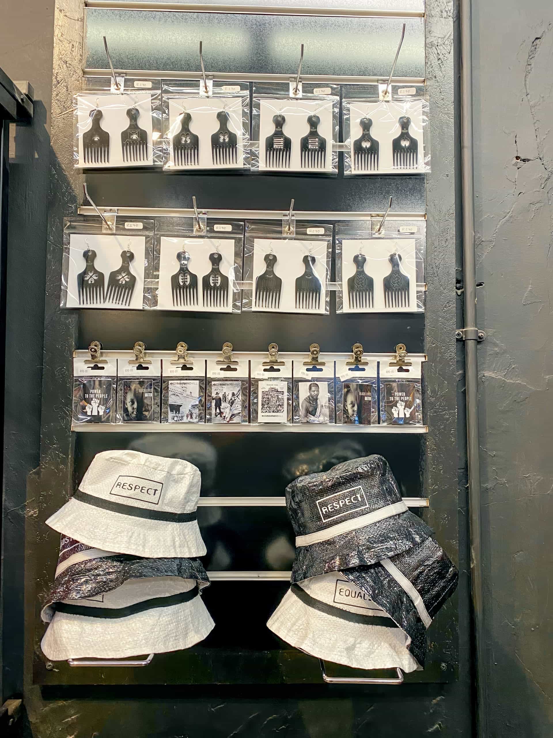 A black display wall features metal afro combs and black-and-white prints of people attached with clips. Two hats labelled 