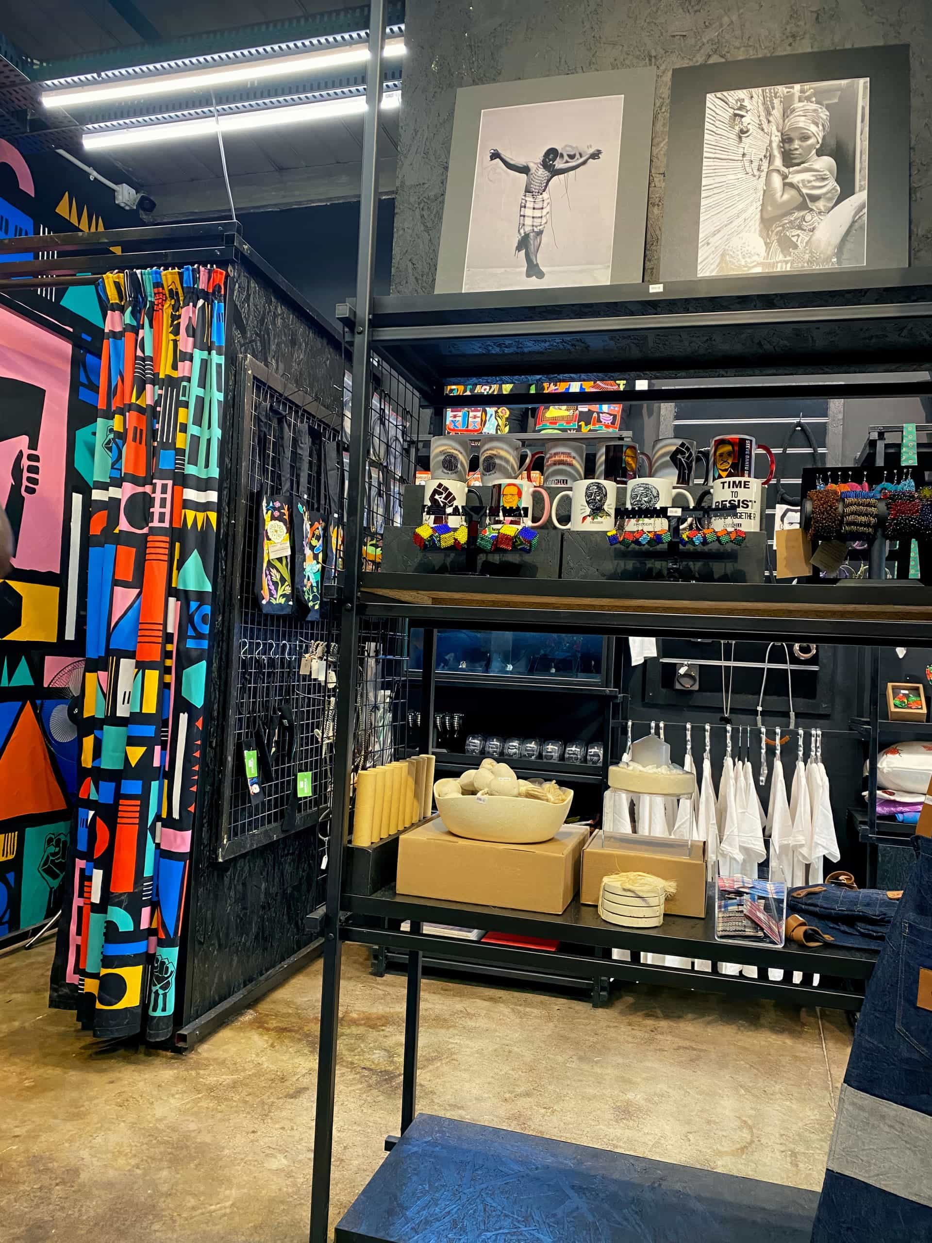 A colourful shop display features vibrant patterned fabrics, art prints of children on a grey wall, mugs with bold designs, beaded jewellery, white aprons, stacked boxes, and shelves with folded textiles. The store has an artistic, modern, and eclectic style.