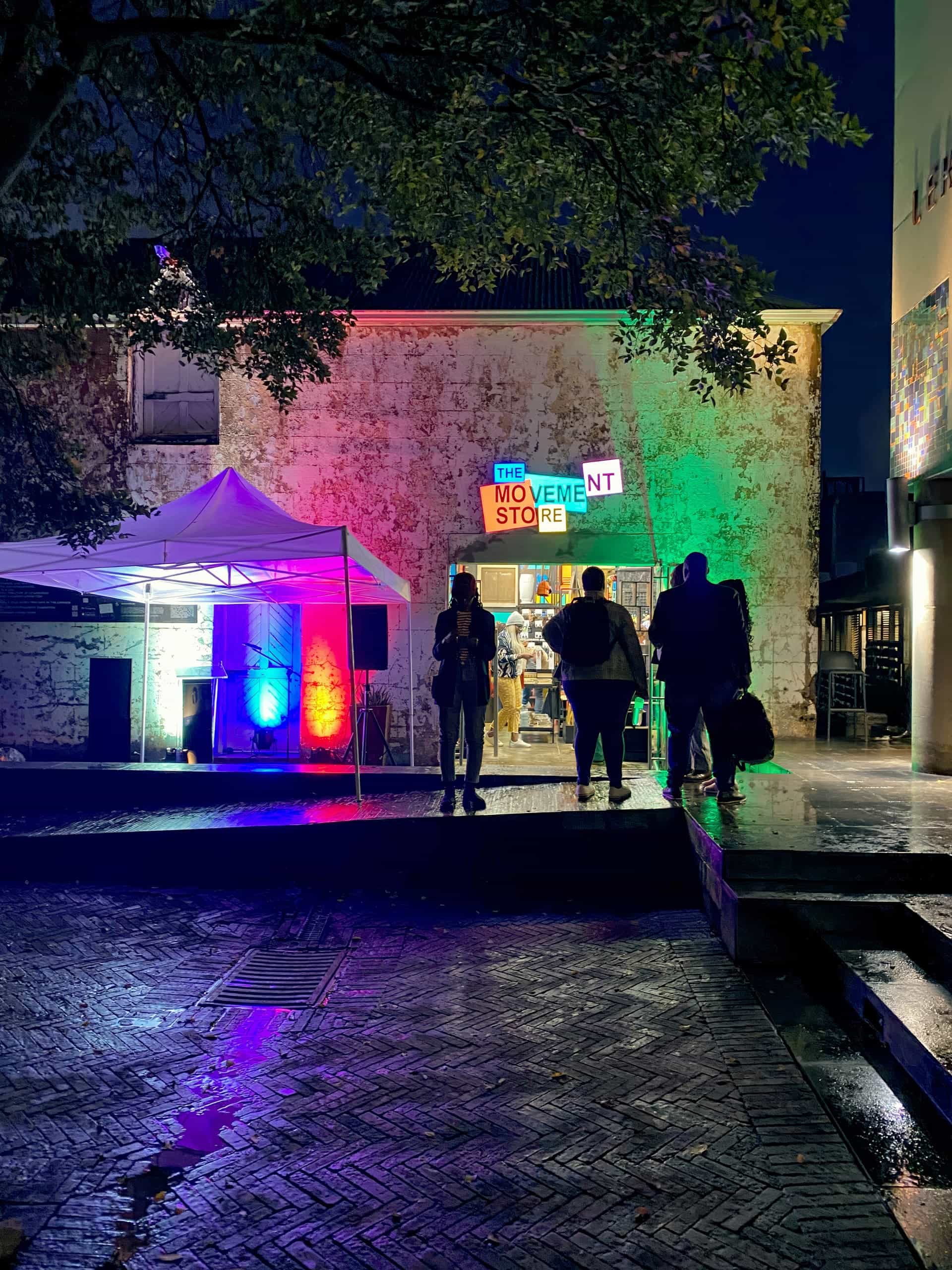 People stand in front of The Movement Shop at night, illuminated by colourful lights (red, green, blue). A white marquee and speakers are set up nearby. Wet pavement and leafy tree branches frame the lively, urban outdoor scene.