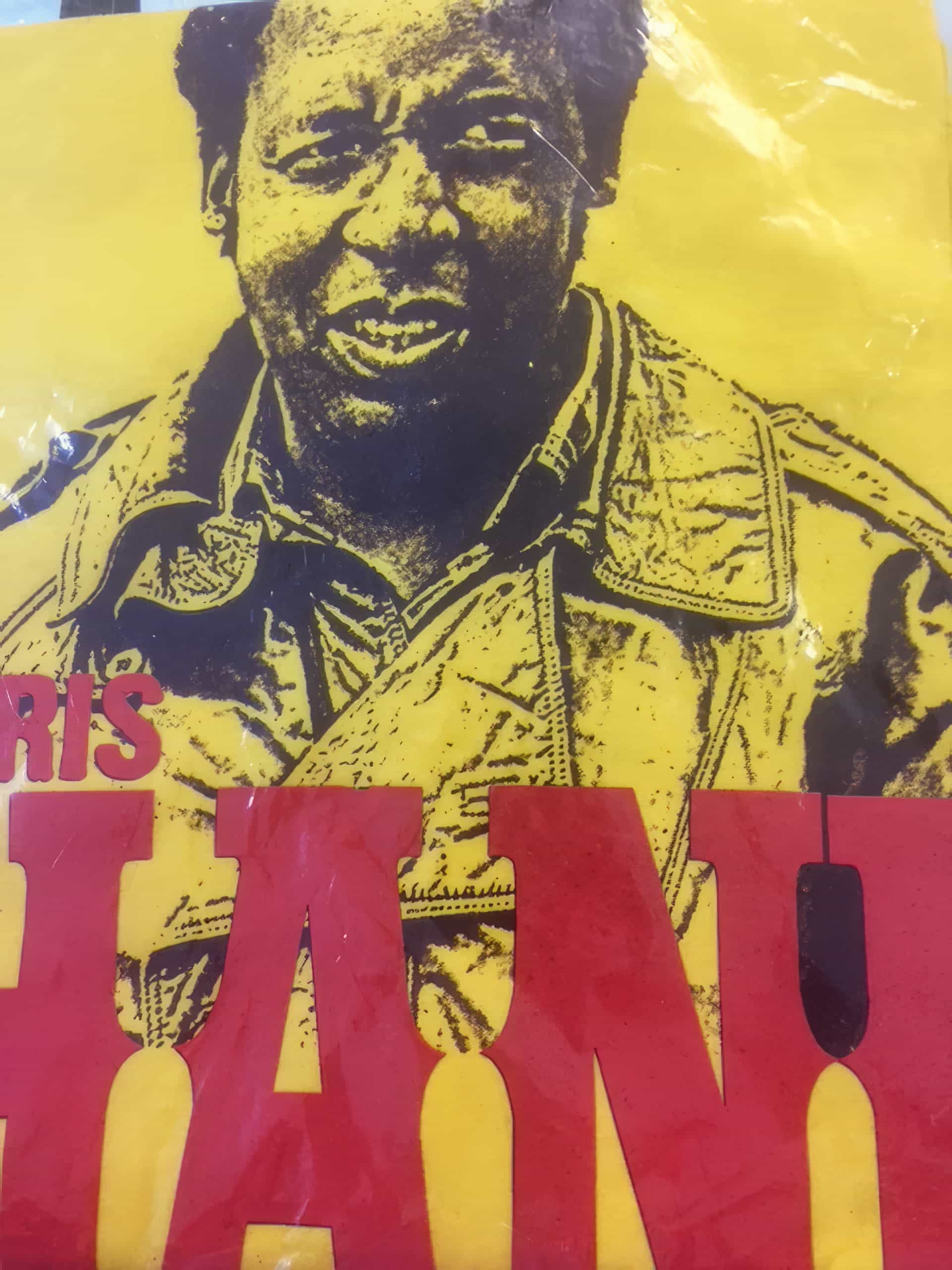 A yellow poster shows a black-and-white portrait of a person wearing a jacket, smiling slightly. Large red block letters partially obscure the lower part of the image. The design has a bold, graphic look.