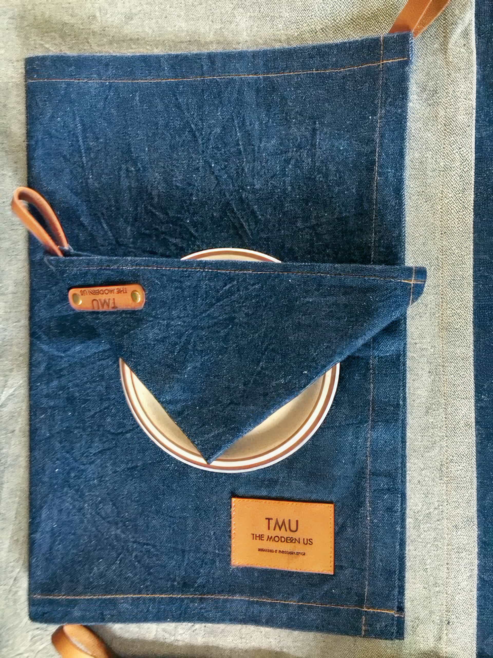 A folded dark blue denim placemat with brown leather accents and a label reading “TMU THE MODERN US.” A white plate with a brown rim is partially tucked into a triangular pocket on the placemat. The surface below is a lighter fabric.