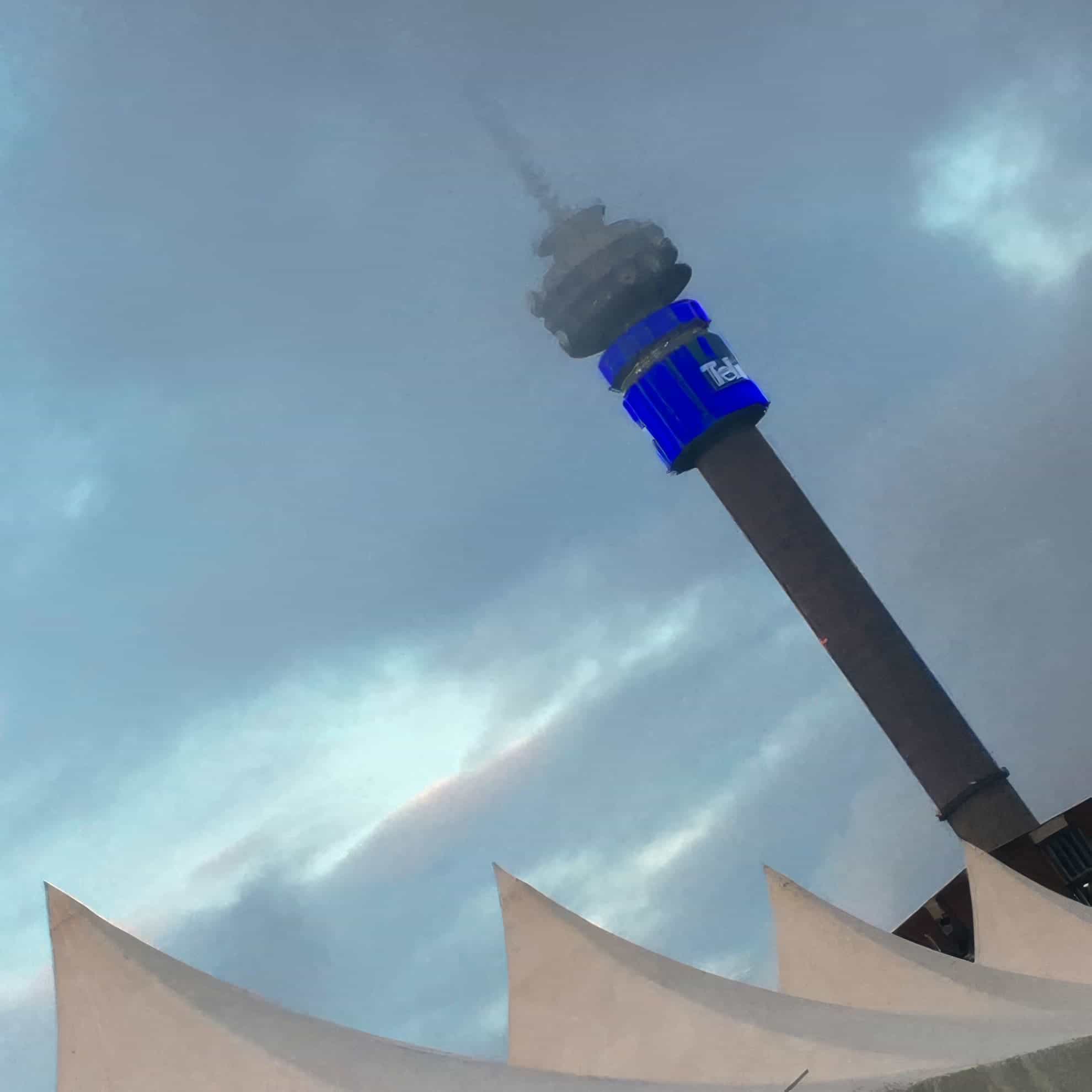 A tall communications tower rises into a cloudy sky, partly shrouded in mist. A blue illuminated sign wraps around the tower near the top. In the foreground, sharp white peaks of a tent-like structure are visible. The atmosphere appears moody and overcast.
