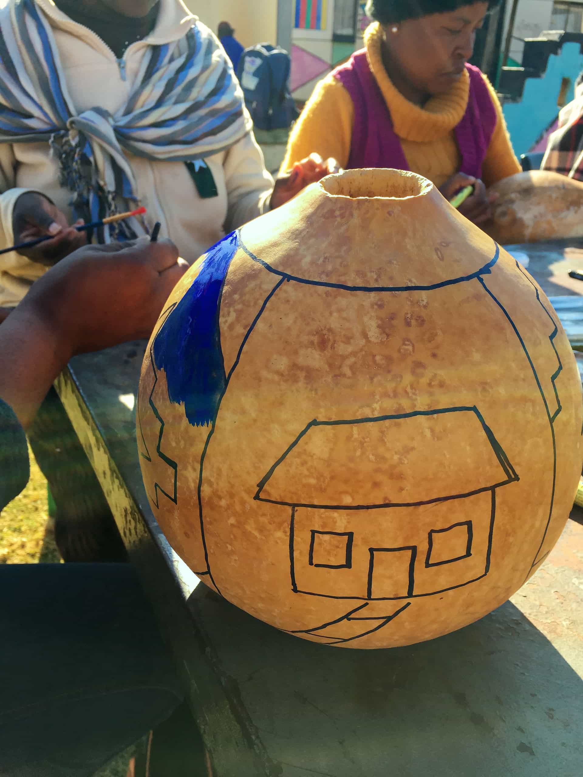 A close-up of a dried gourd with simple house outlines drawn in black marker; a person is painting part of the design blue. In the background, two people sit at a table, focused on their own work, wearing warm clothing and scarves.