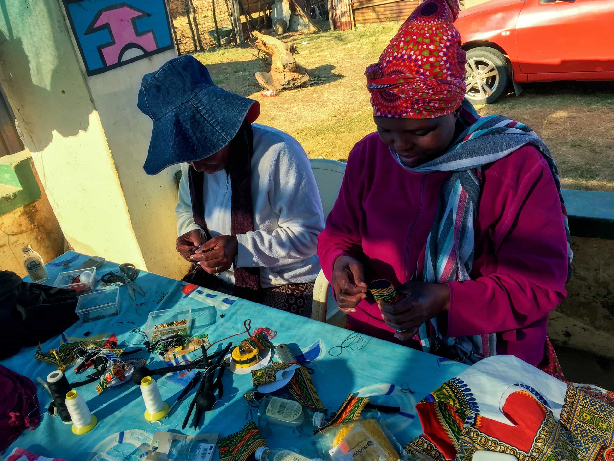 Two women sit at a table covered with colourful beads, thread, and craft supplies, working on beaded jewellery. Both wear head coverings and warm clothes. Sunlight casts shadows across the table. In the background, there’s a red car and a wall with a painted mural.