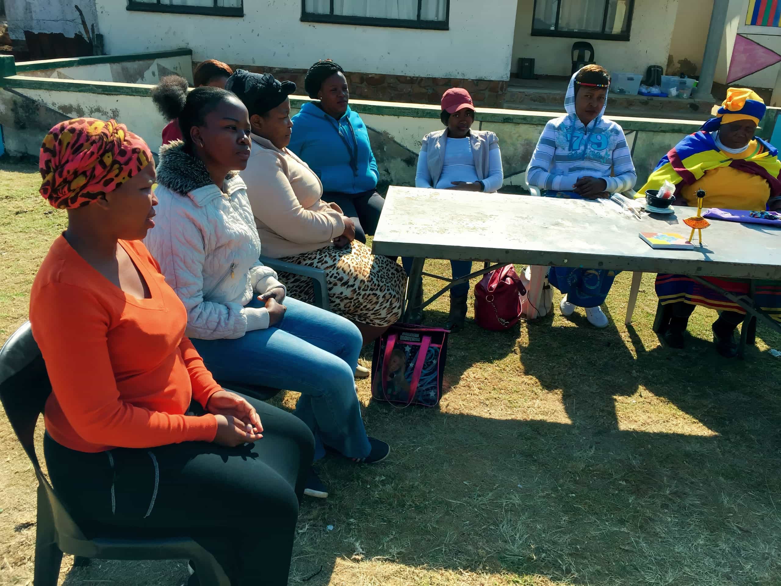 A group of women sit outdoors on plastic chairs around a rectangular table. They appear to be engaged in a discussion or meeting. Some women wear headscarves and brightly coloured clothing. The scene is sunny, with a building and windows in the background.