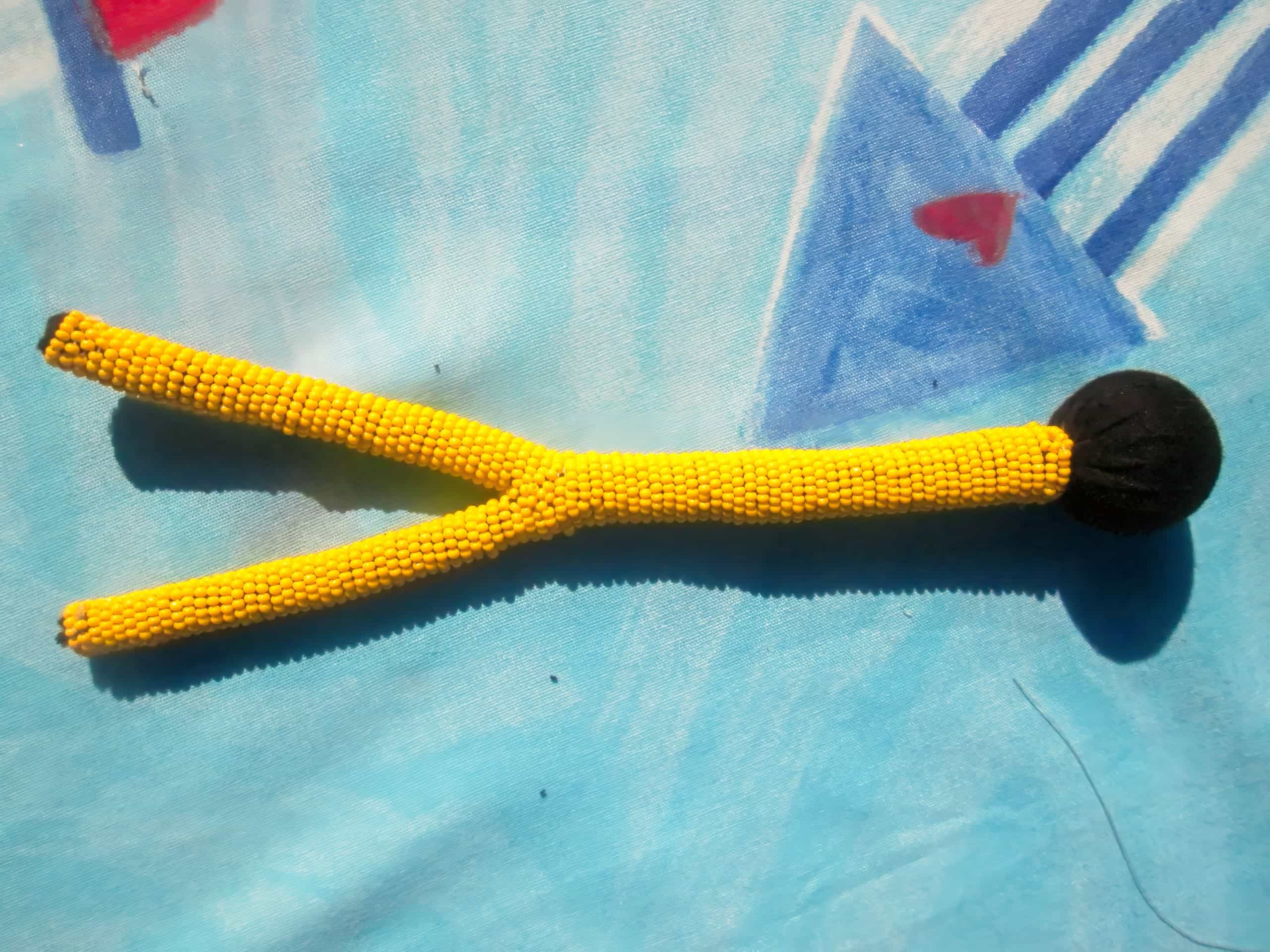A yellow Y-shaped object made of small beads with a black round end rests on a blue surface decorated with abstract triangle and heart shapes. Shadows are visible, and the background has a soft, textured appearance.