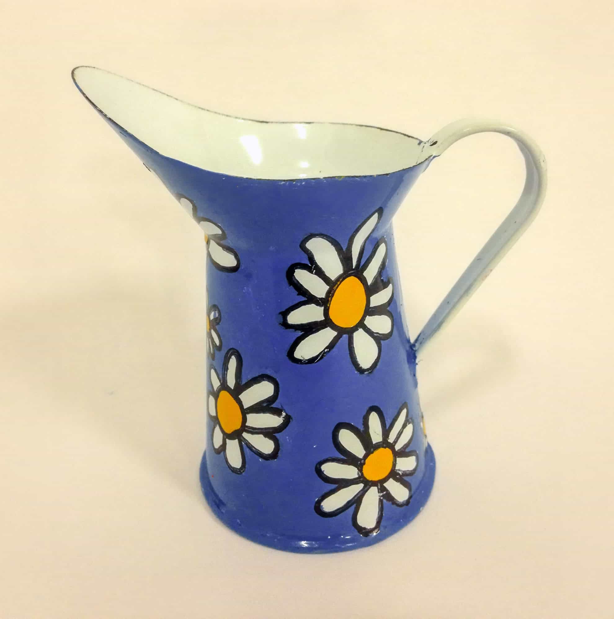 A blue metal jug with a side handle, decorated with white and yellow daisy flowers outlined in black. The inside of the jug is white, and the background is plain and light-coloured.