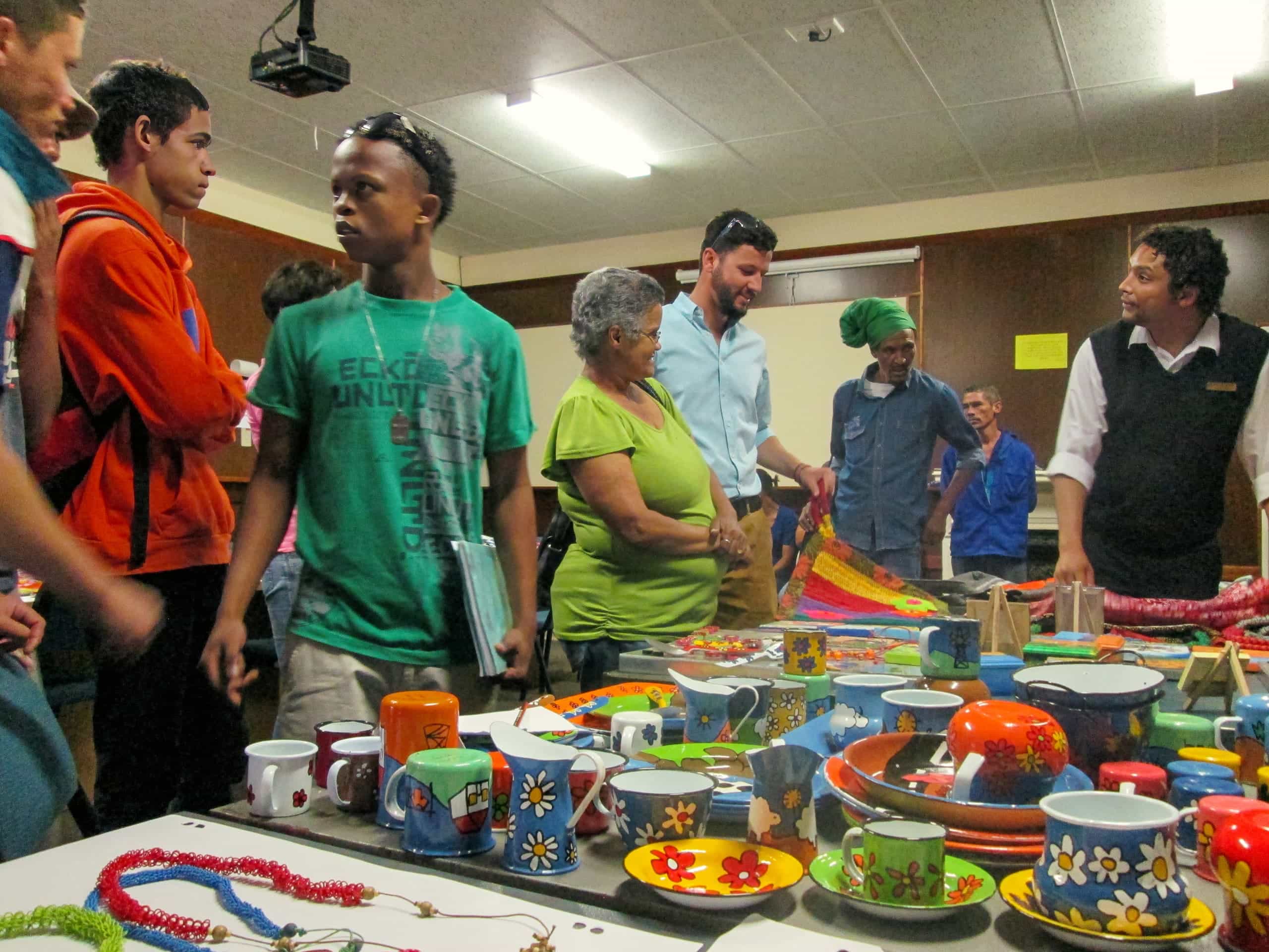 A diverse group of people stands and talks around tables covered with colourful, hand-painted pottery, mugs, bowls, and jewellery at an indoor craft fair. The room has brown walls, fluorescent lights, and a warm, lively atmosphere.