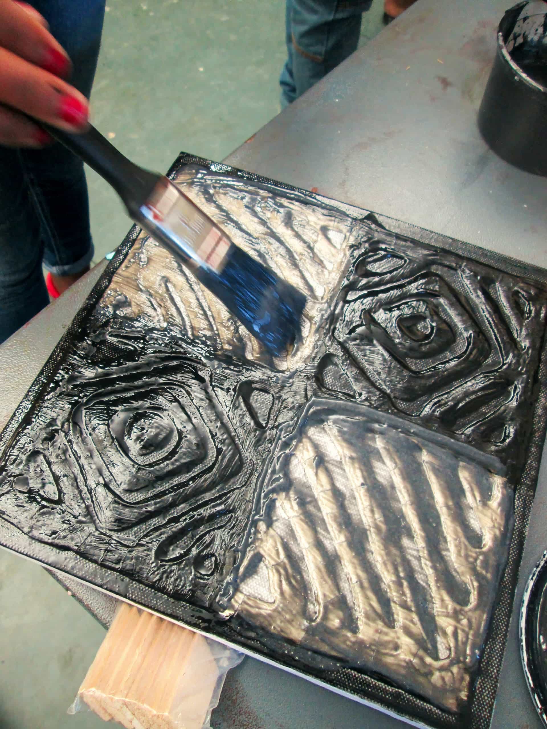 A hand with red nail varnish uses a paintbrush to apply black paint onto a textured, square tile with geometric patterns. The tile is on a work surface, and another person stands in the background.