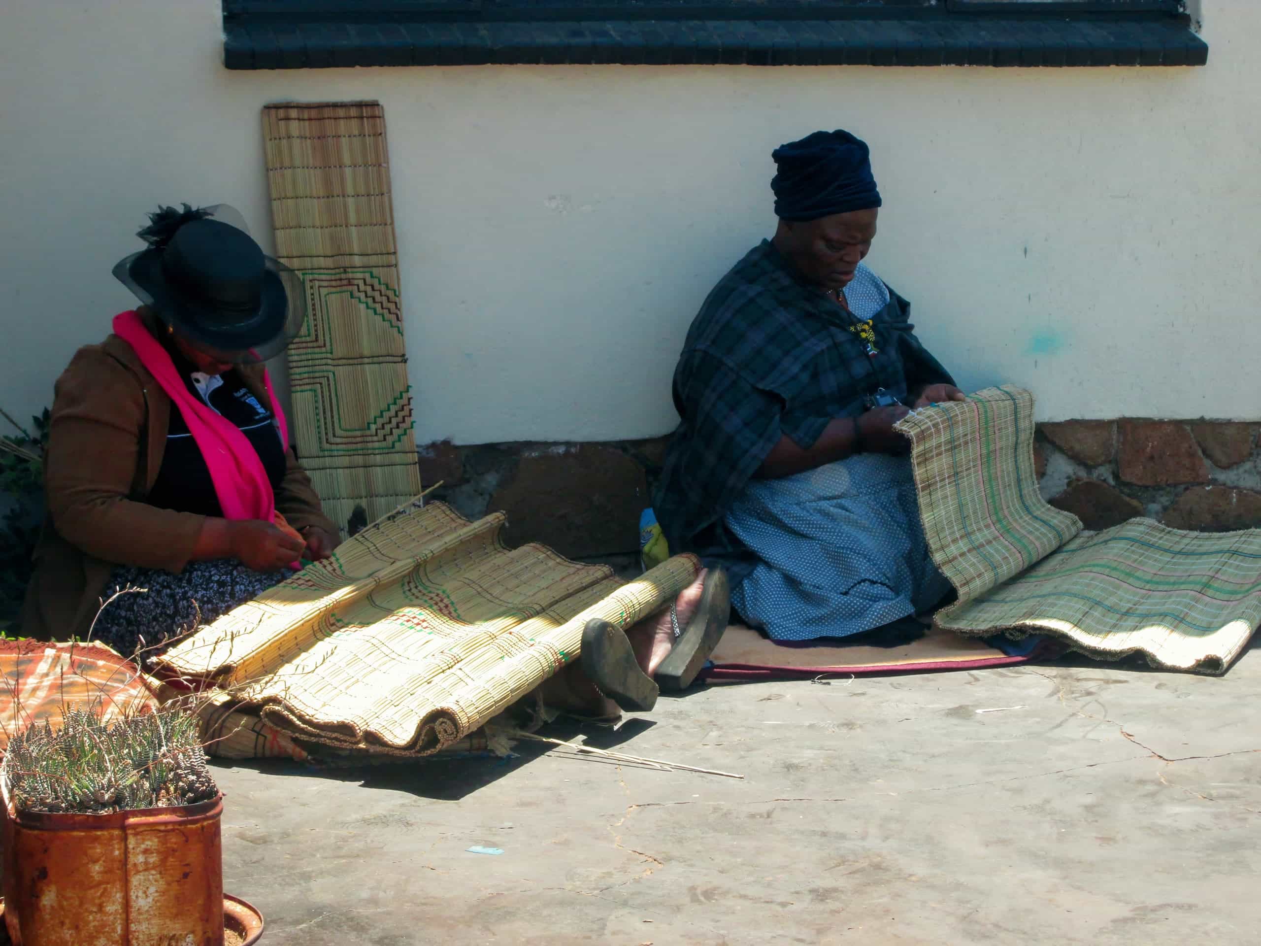 Two women sit on the ground outside, weaving traditional mats from straw. One wears a hat and pink scarf, the other a blue headscarf and checked shawl. Rolled and finished mats are beside them, with a small potted plant in the lower left corner.