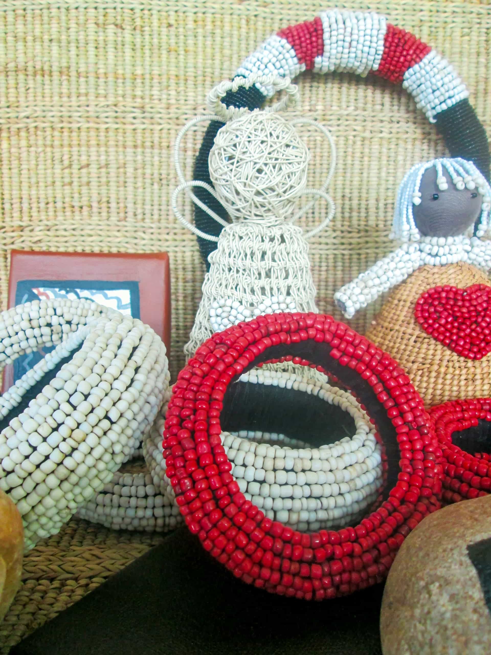 Handcrafted beadwork including white and red bracelets, a wire angel, and a doll with a stitched red heart, are displayed against a woven background. The items are arranged closely together, showcasing their textures and intricate details.