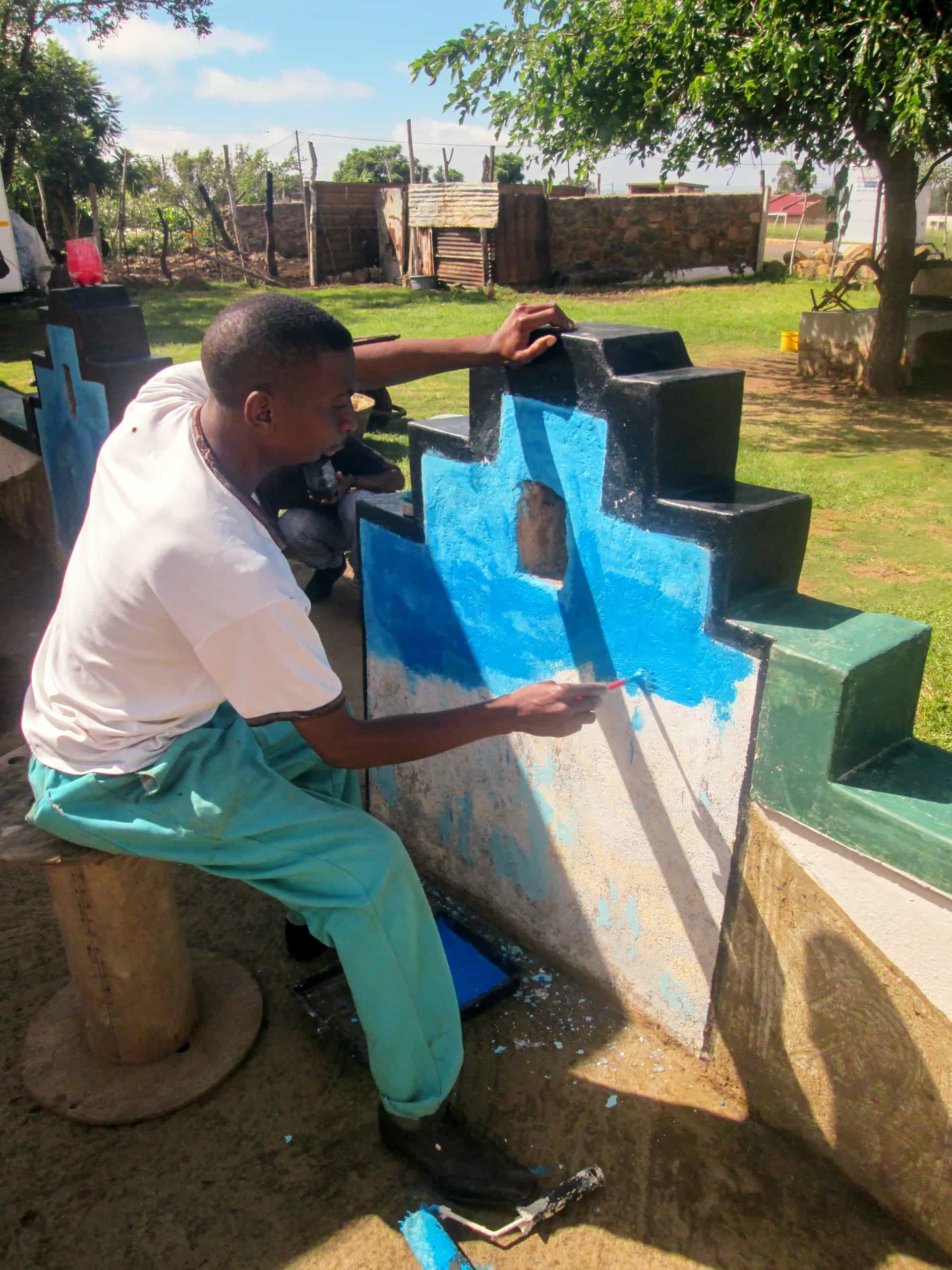 A man sits on a stool outdoors, painting a geometric concrete wall bright blue. He wears a white shirt and teal trousers and uses a small brush. Green grass, trees, and wooden fencing are visible in the sunny background.