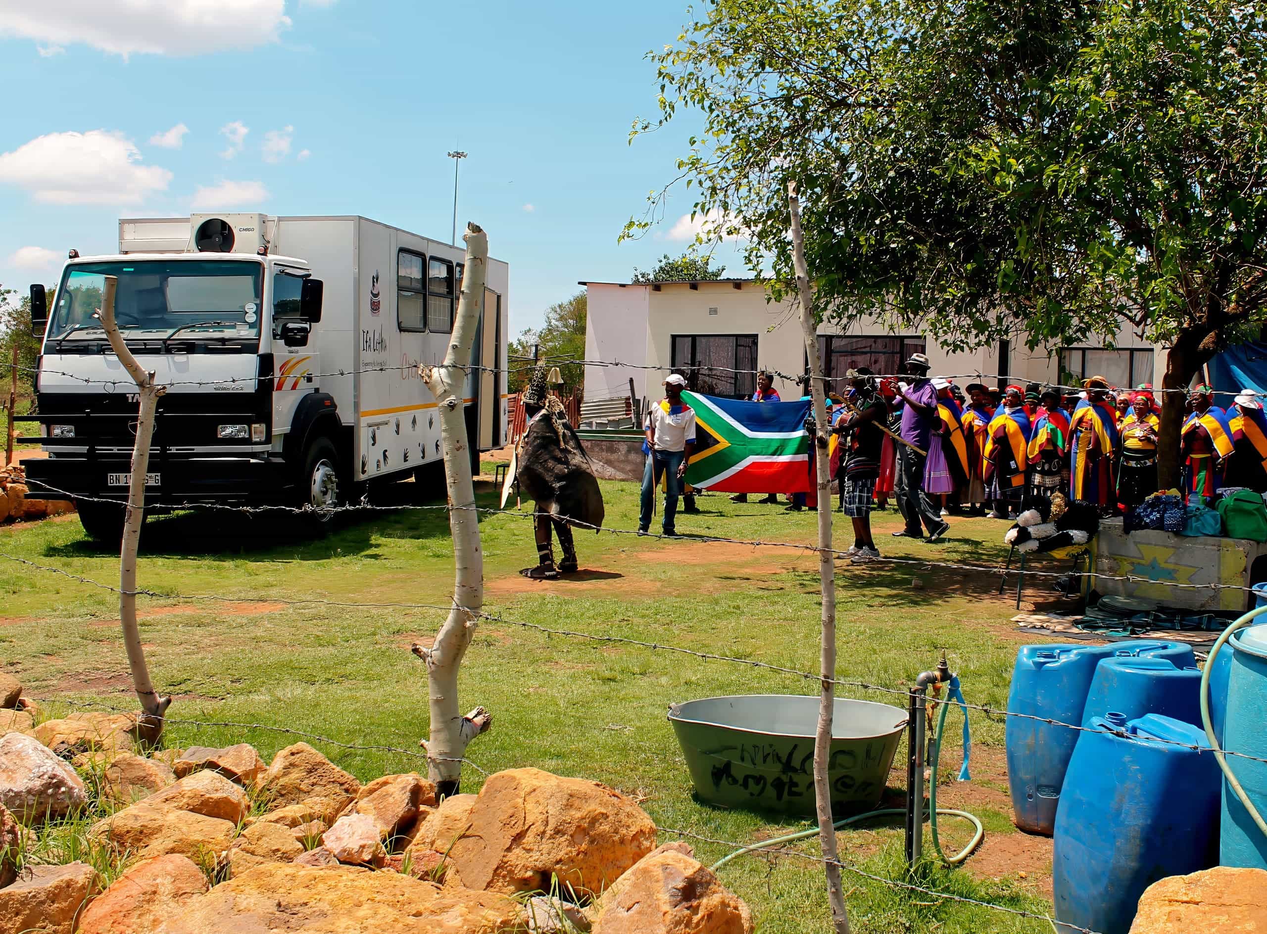 A group of children in colourful uniforms stand near a building and South African flag. In the foreground, blue water barrels, a metal tub, and rocks are visible. A large white lorry and a person in traditional attire appear nearby on grassy ground.