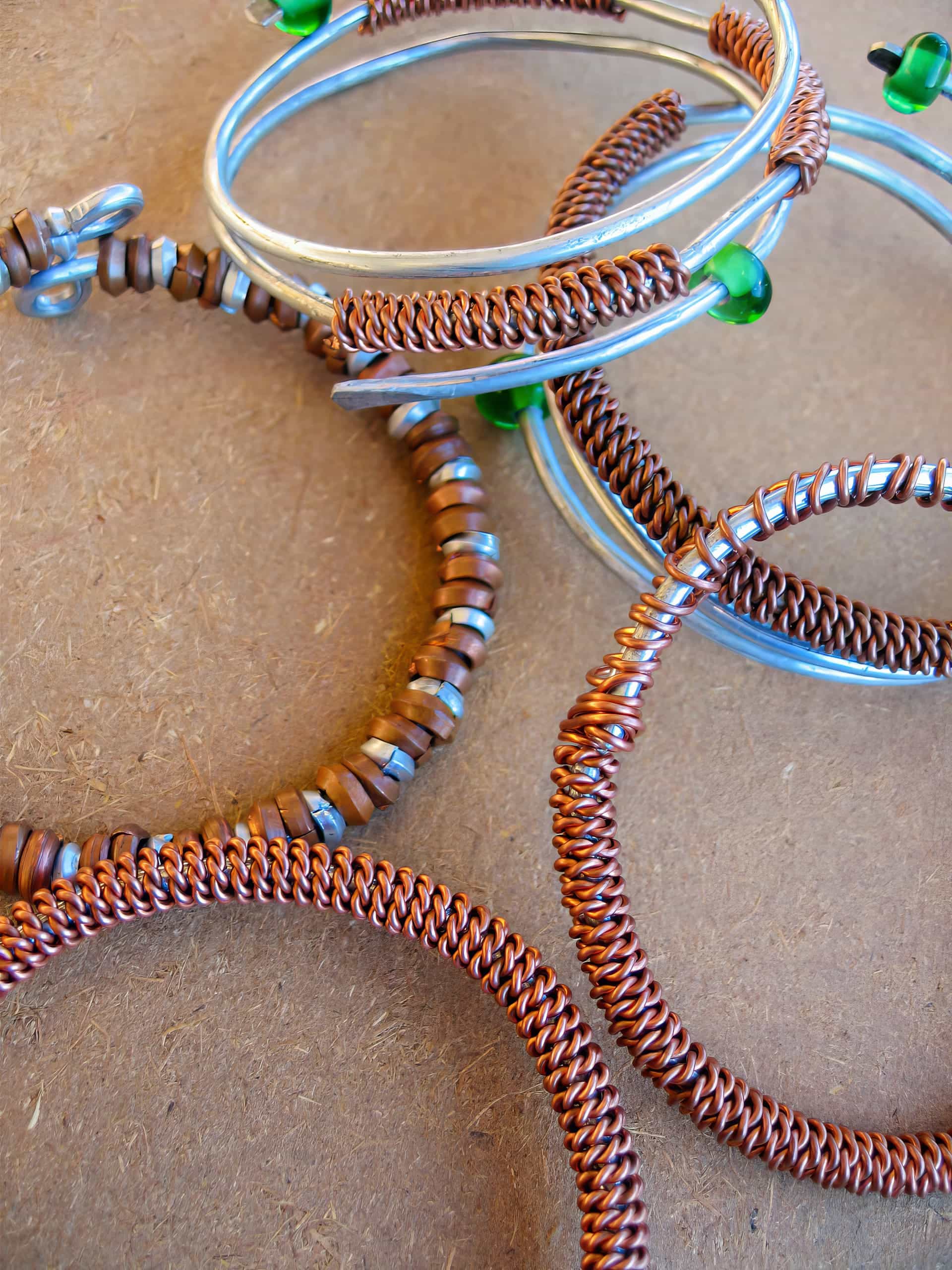 Several handmade wire bracelets rest on a brown surface. Some are copper-coloured with intricate twisted designs, while others are silver with green beads and tan circular beads threaded along the wire. The bracelets overlap and create a textured, artistic arrangement.