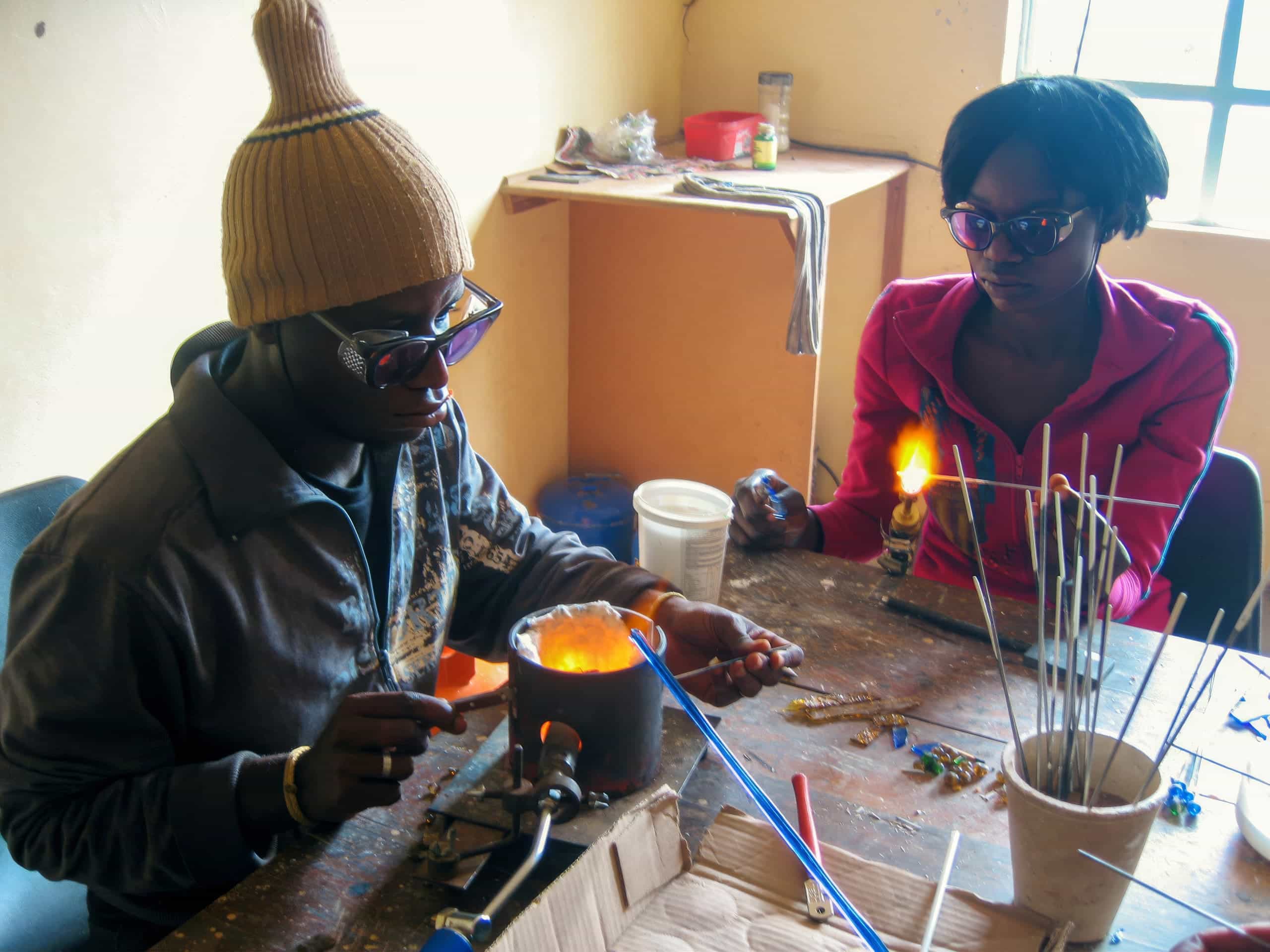Two people wearing sunglasses work with hot glass at a cluttered table. One uses tools over a small furnace; the other holds a rod with glowing molten glass. The room is sunlit, with shelves, sticks, and glasswork supplies scattered about.