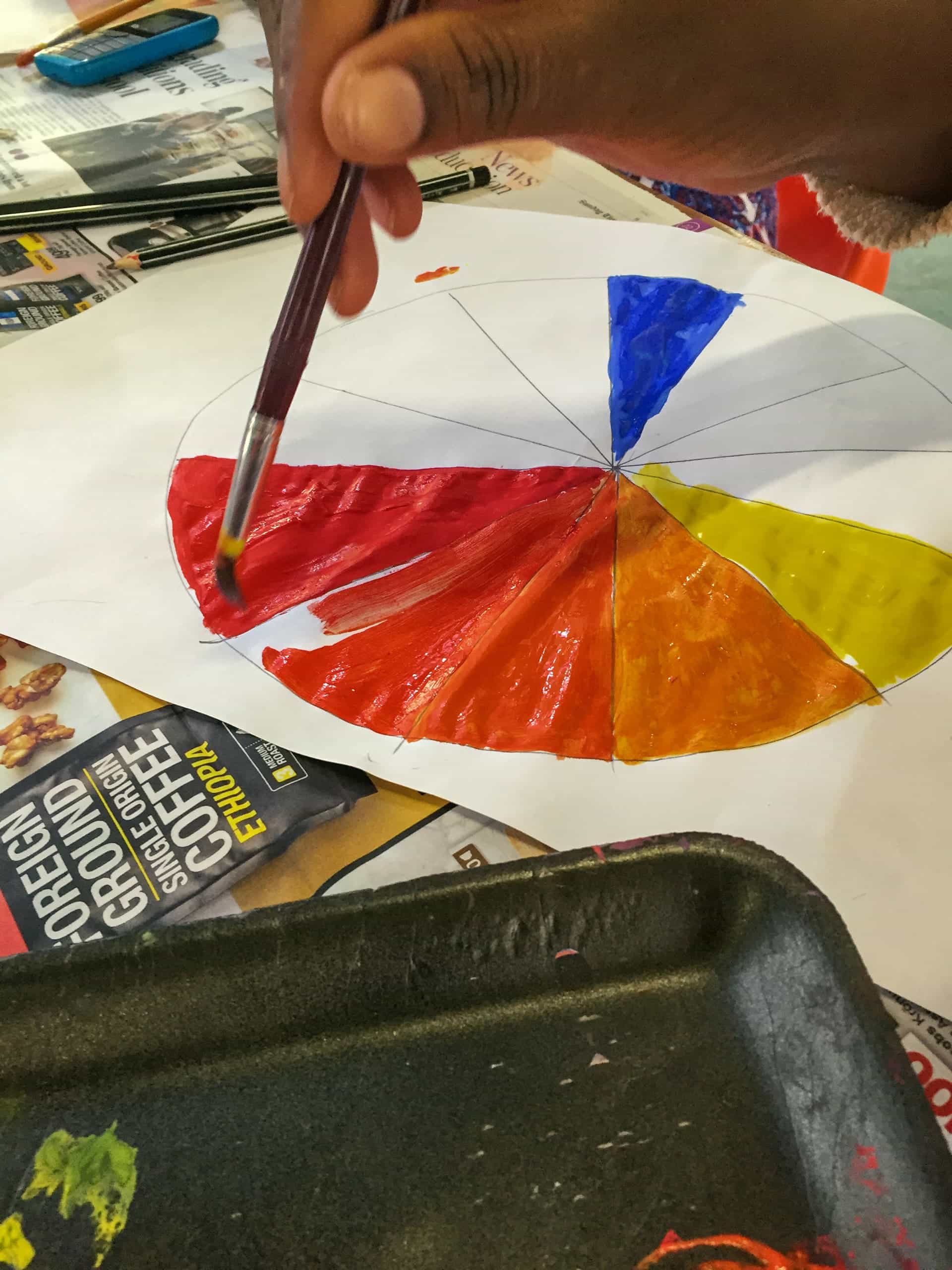 A hand holds a paintbrush, painting sections of a colour wheel on white paper. The wheel includes red, yellow, blue, and orange. Nearby are newspapers, a black tray with paint, and a coffee advertisement on the table.