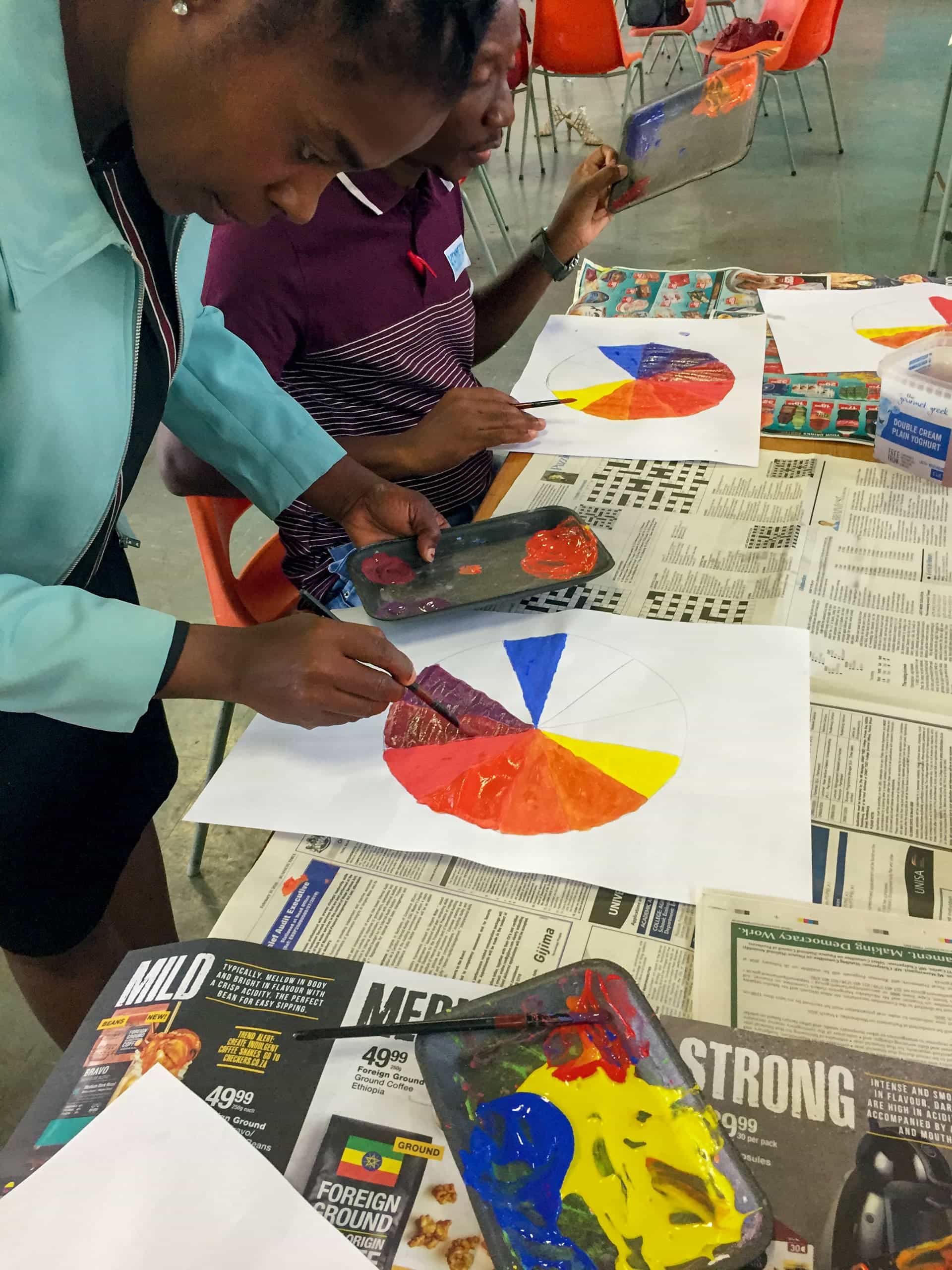 Two people sit at a table covered with newspapers, painting colour wheels on white paper using brushes and paint palettes. One person in the foreground is focused on adding purple paint, whilst the other mixes colours. Orange chairs are in the background.